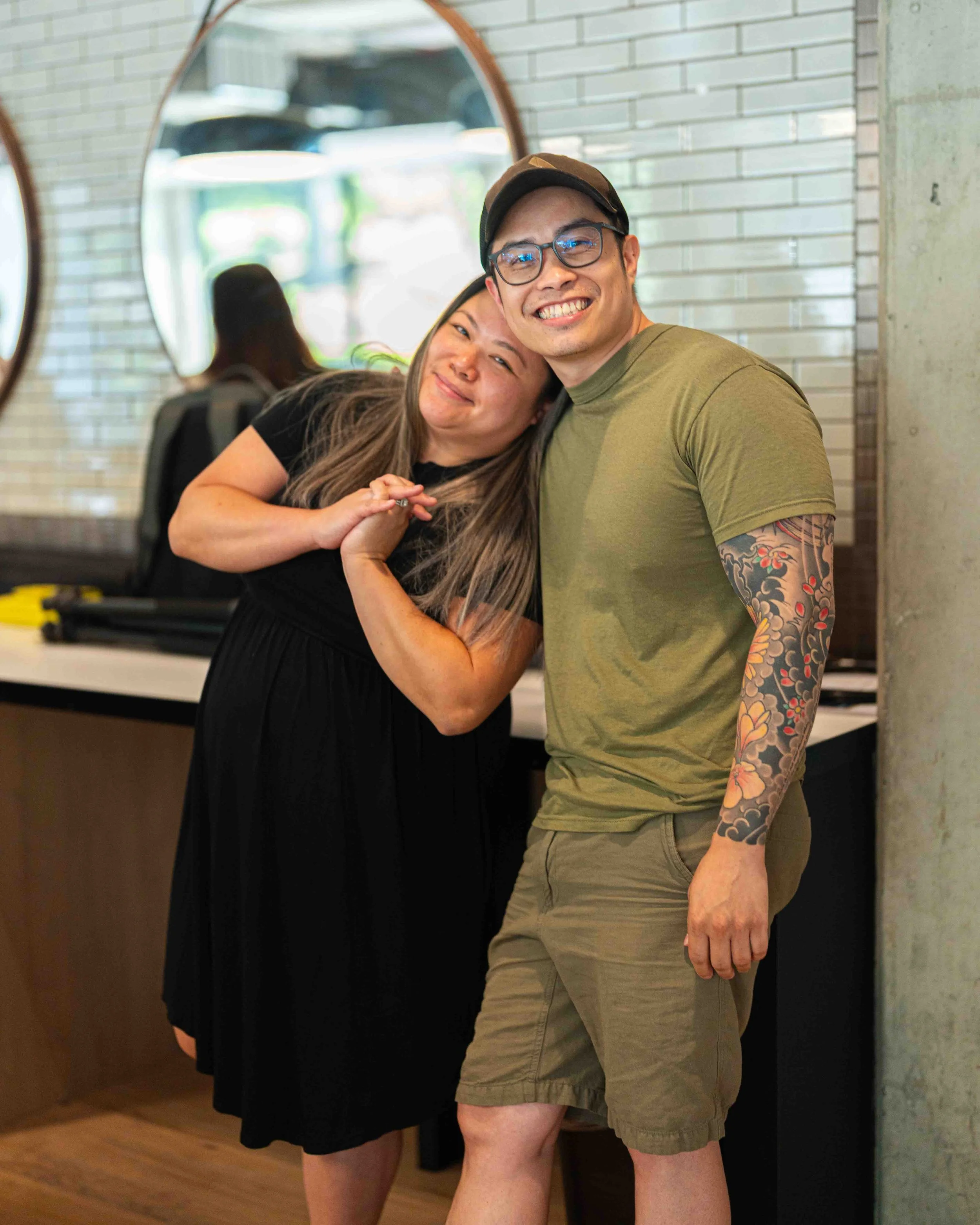 Two smiling people, a woman with long hair and a man with glasses and a tattooed arm, standing close together indoors near a mirror with a brick wall behind them, both with casual clothing and cheerful expressions.