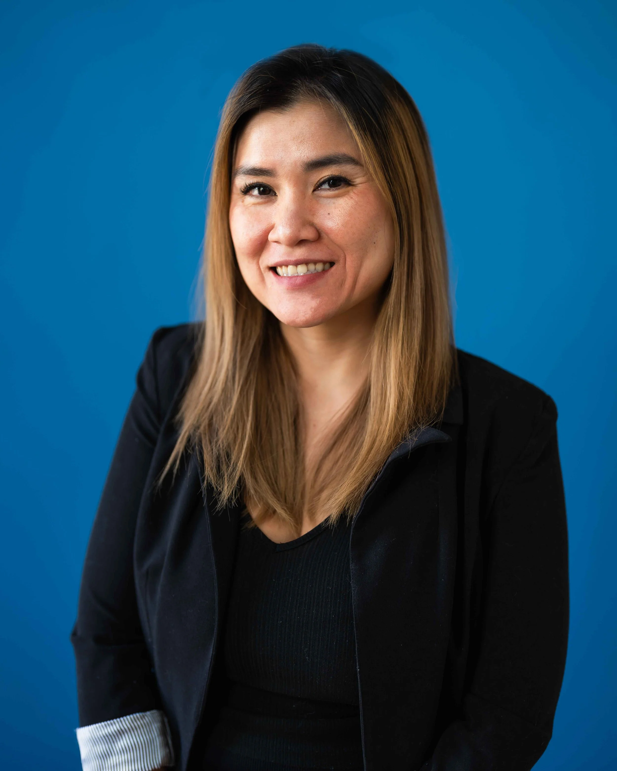 portrait of a woman with long hair, smiling, wearing a black blazer and a black top, against a blue background