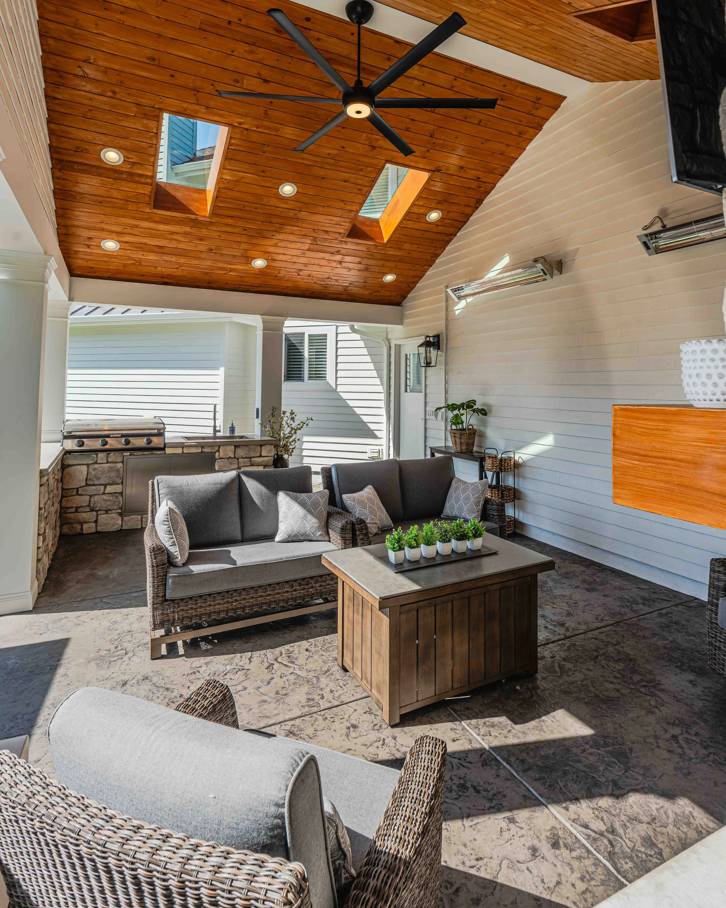 A patio area with wicker chairs, a wood table with small potted plants, a sectional sofa, a stone outdoor kitchen grill, white siding house walls, and a wooden ceiling with skylights and a black ceiling fan.