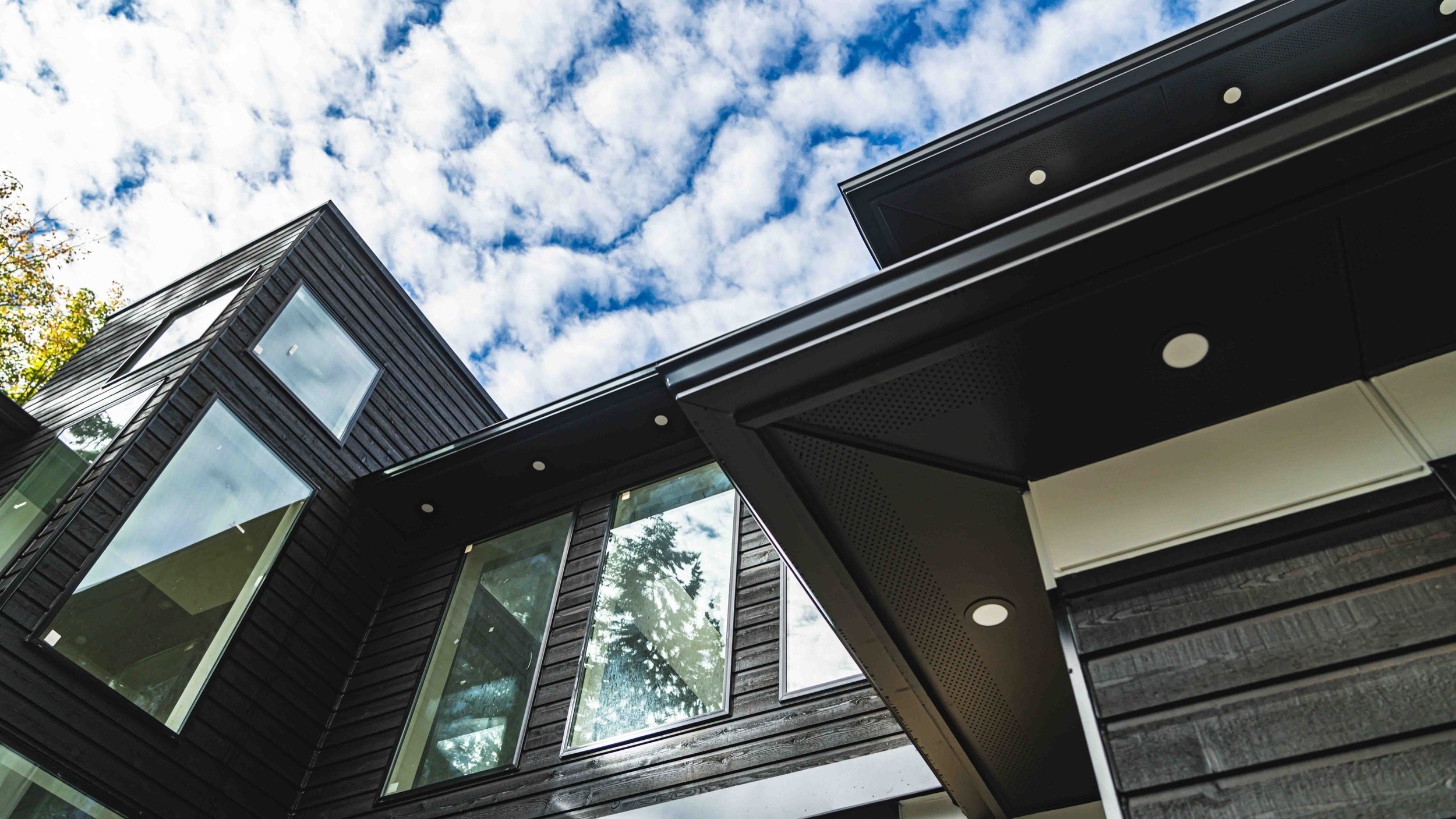 Low-angle view of modern black wooden and glass house exterior with bright blue sky and white clouds overhead.