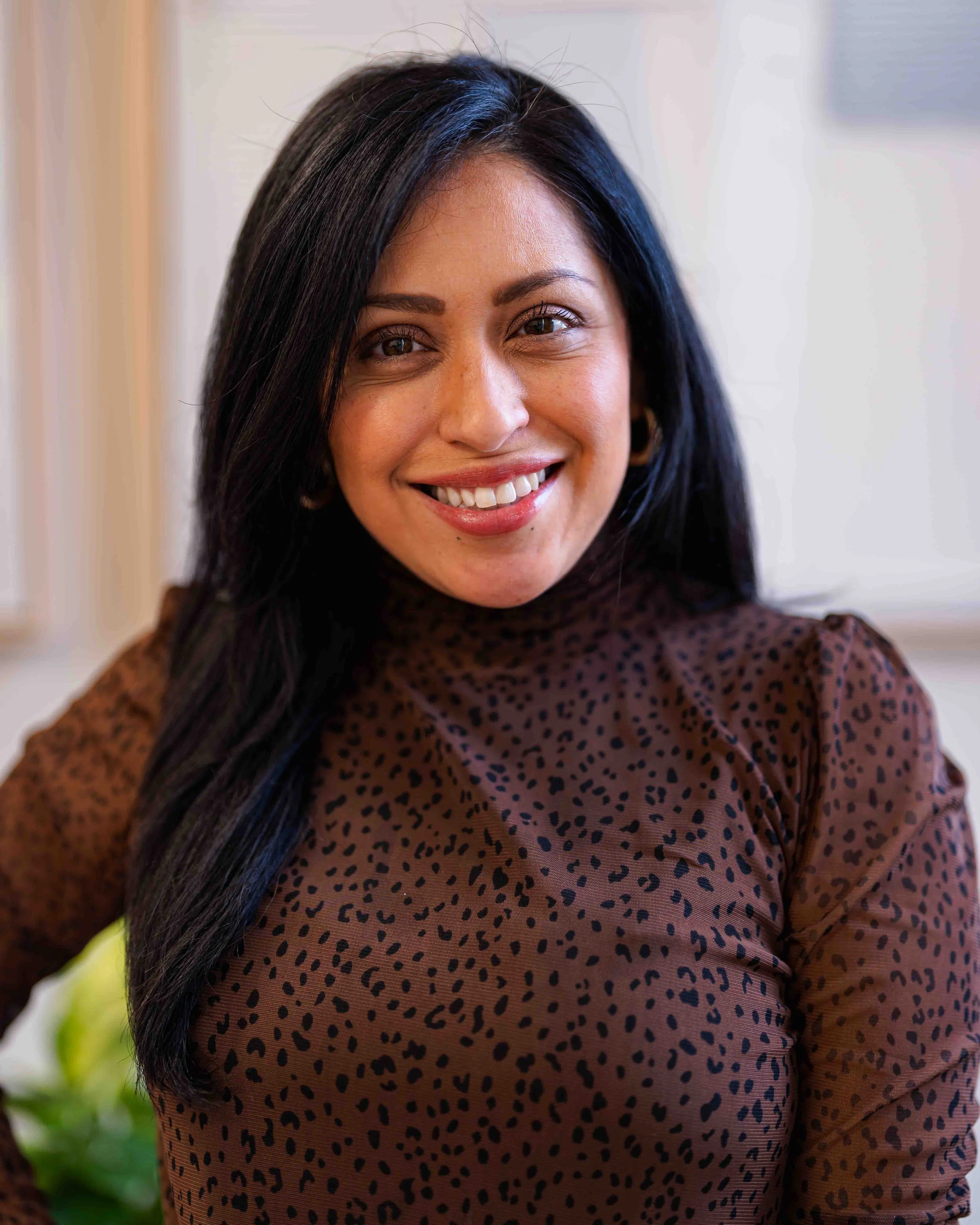 Close-up portrait of a woman with long dark hair, smiling and wearing a brown leopard print top, indoors with blurred background.