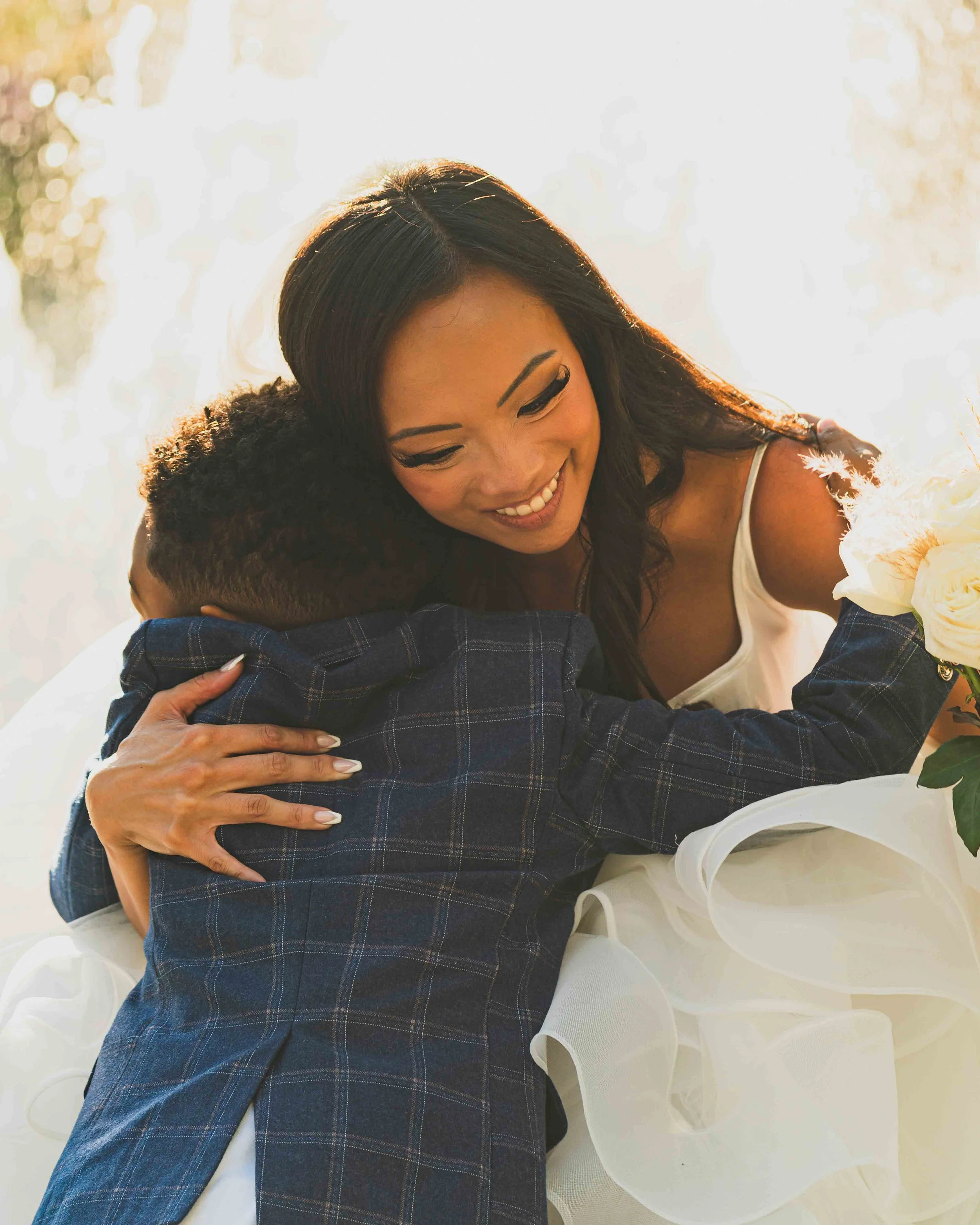A woman hugging a young boy at an outdoor event, both smiling. The woman has long dark hair and is wearing a white top. The boy has short hair and is wearing a blue plaid shirt. The background is blurred with bright sunlight.