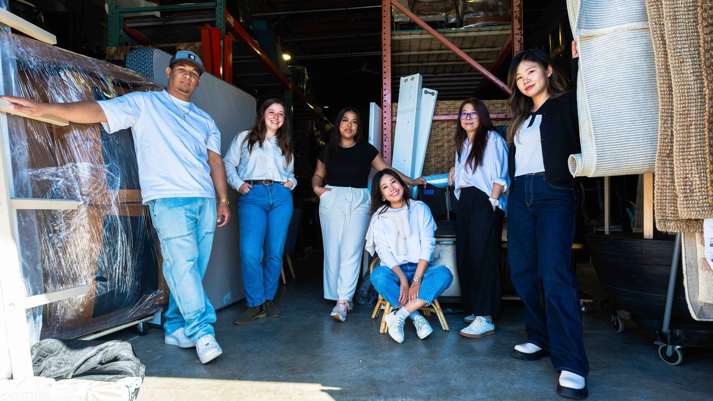 Group of six diverse people standing and sitting inside a warehouse, smiling and posing for the camera.