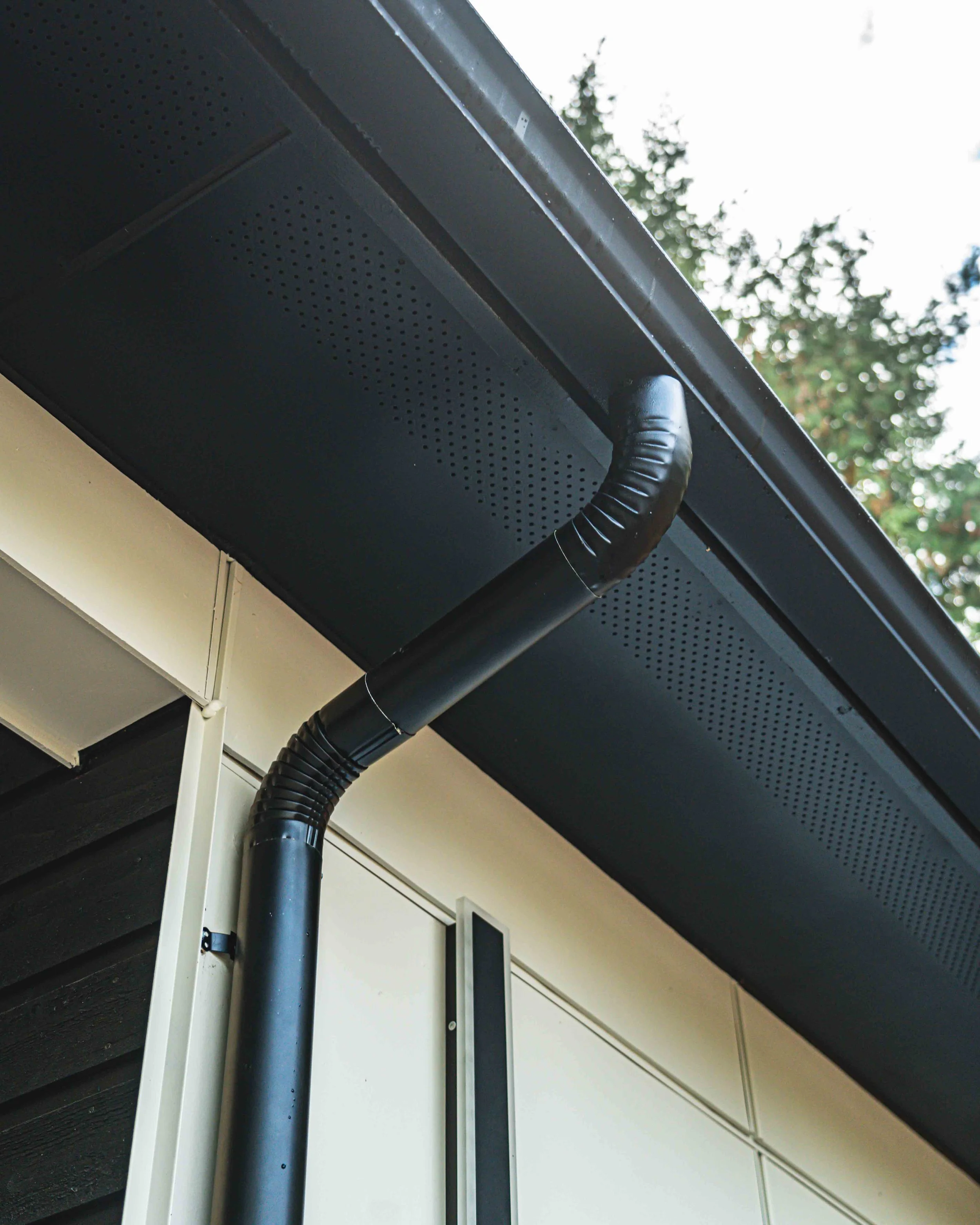 Close-up of black rain gutters and downspout on the side of a building, with some trees and sky in the background.