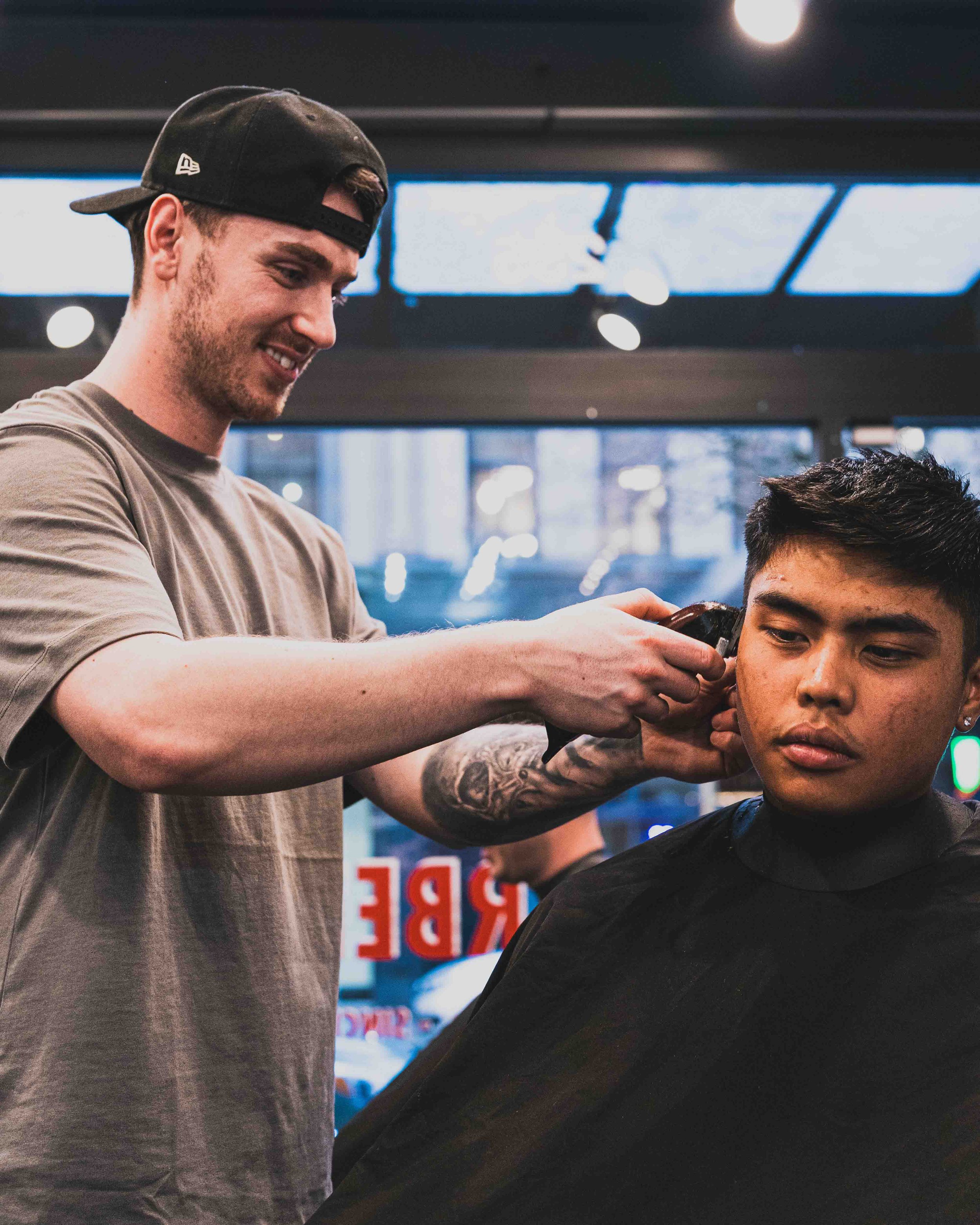 A barbergiving a haircut to a young man, with the barber smiling and wearing a black cap backward in an indoor space with bright lighting and a large window in the background.