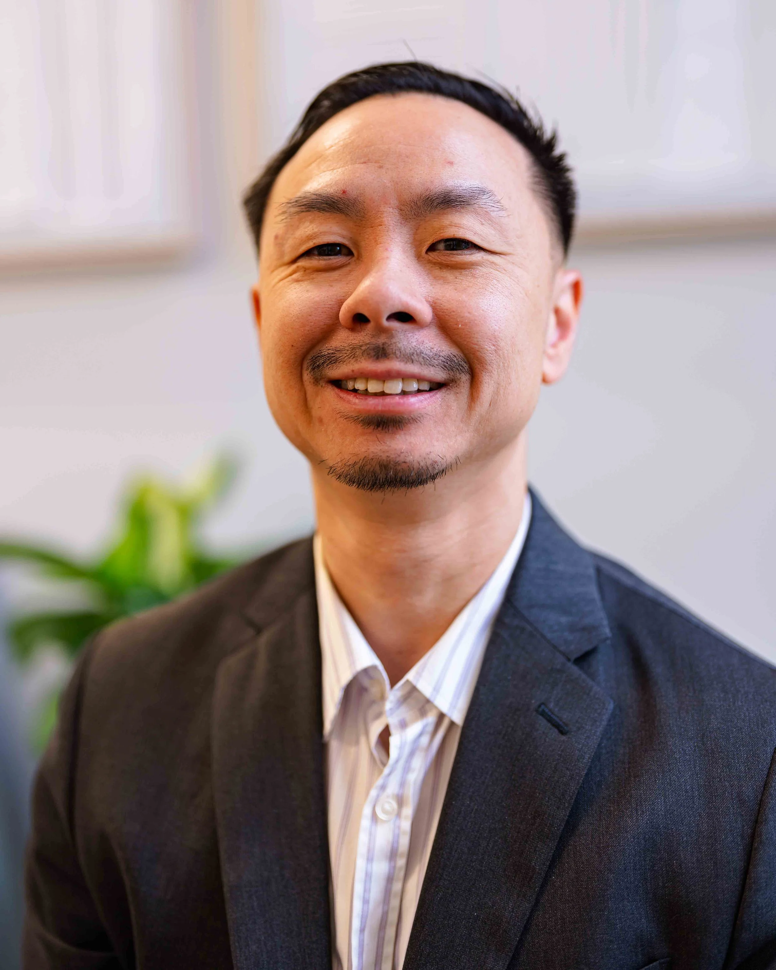 Close-up portrait of smiling middle-aged Asian man in business suit with a striped shirt, indoor setting with blurred green plant in background.