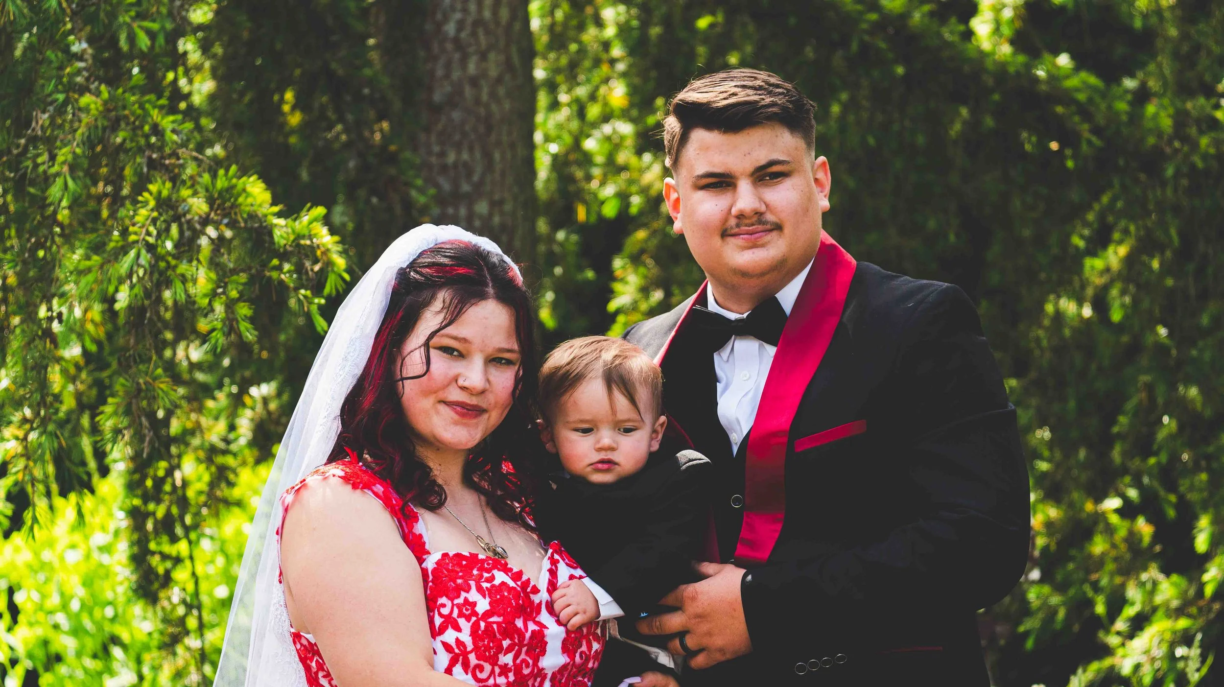 A family of three adults and a young child posing outdoors in a wooded area during daytime. The woman wears a red and white dress and a veil, the man is in a black tuxedo with a red sash, and the child is in a suit. They are standing close together, 