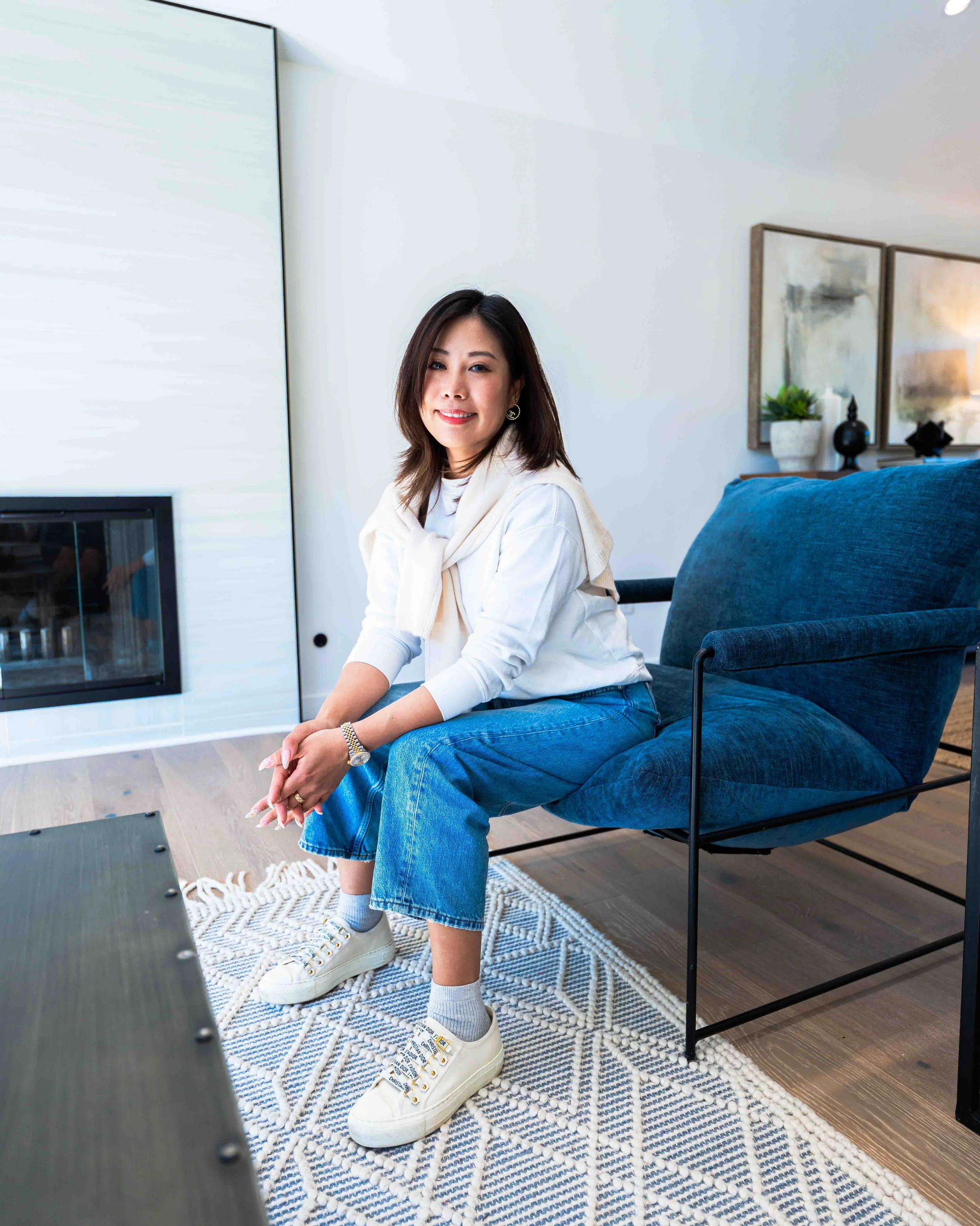 A woman sitting on a chair in a modern living room smiling at the camera. She has medium-length brown hair, is wearing a white long-sleeve top, blue jeans, white sneakers, and has a watch and a bracelet. The living room has a blue armchair, a pattern