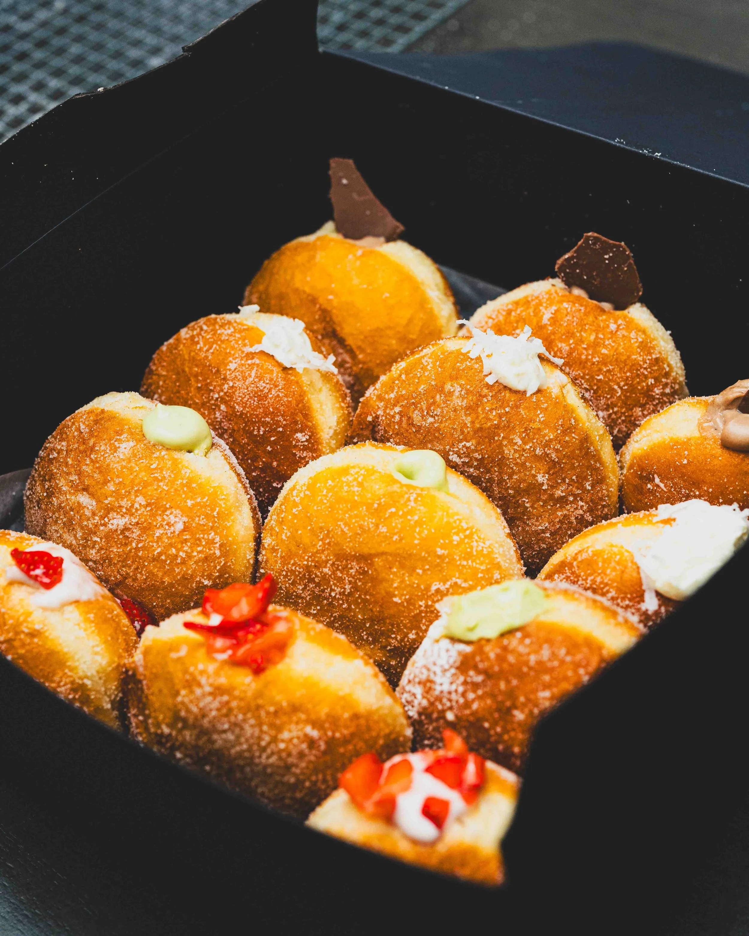 Close-up of a black box filled with assorted colorful desserts, including mini cakes topped with chocolate pieces, fruit, and cream.