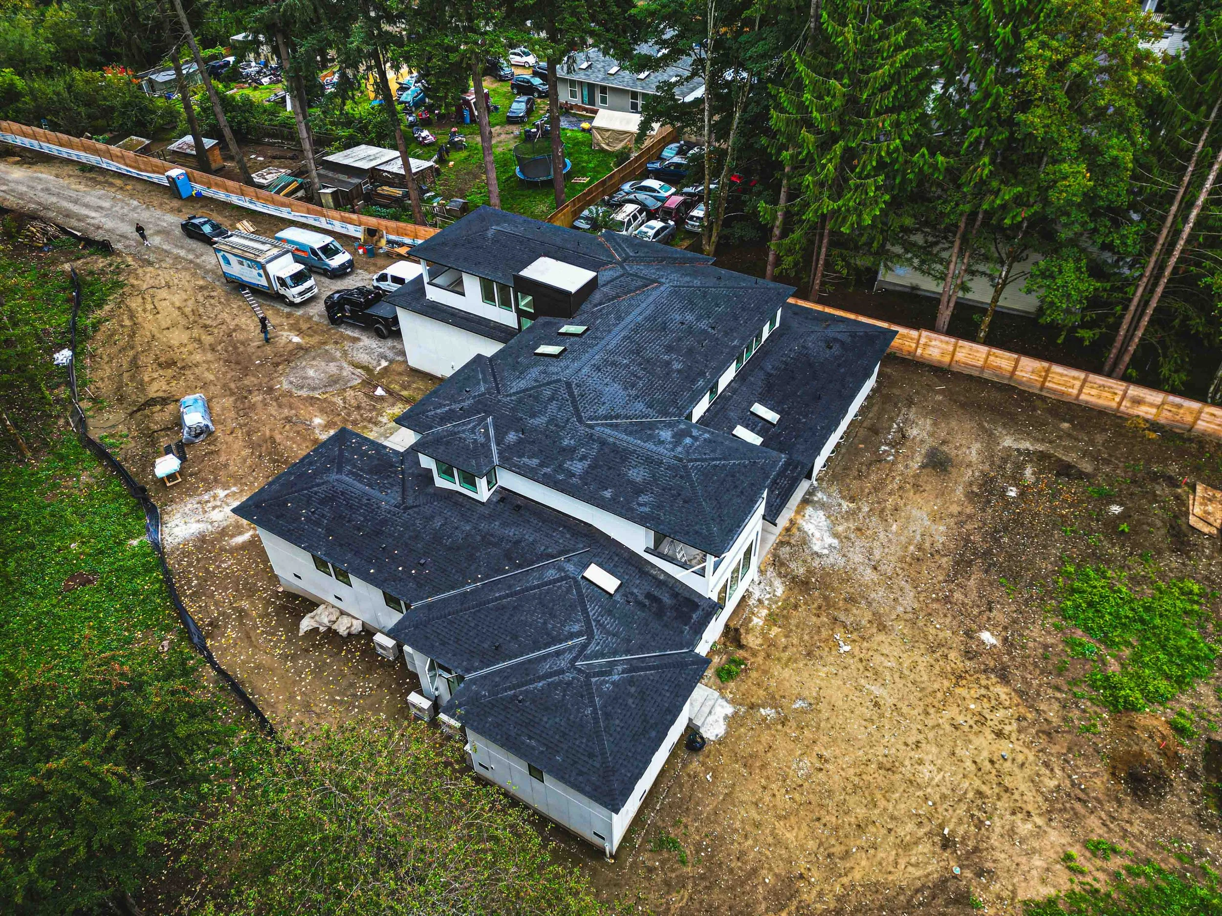 Aerial view of a residential backyard and side yard with cars parked on a dirt driveway, a newly constructed white house, and a fenced-in grassy area with trees and parked vehicles.