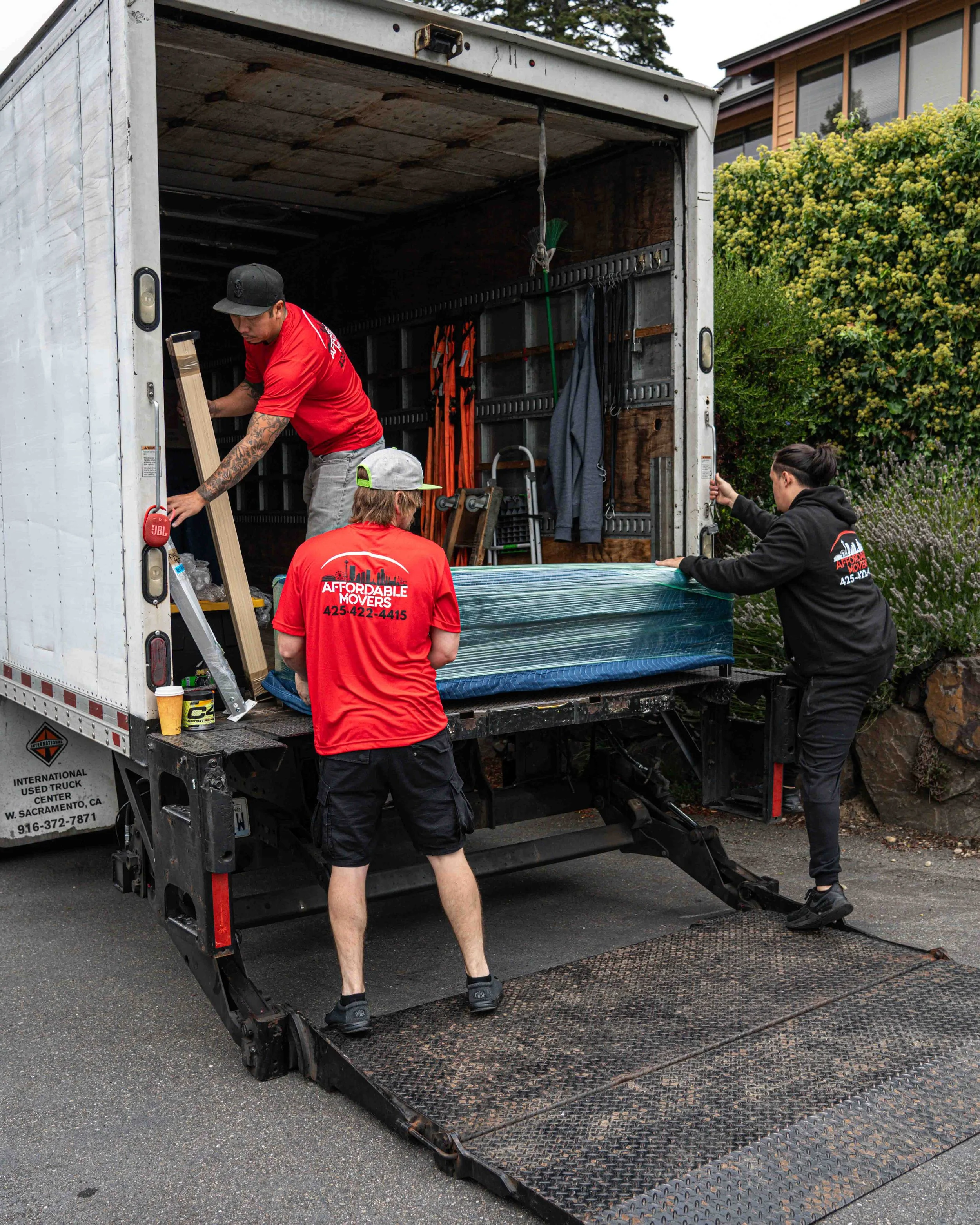 Four people moving a large wrapped object from a truck into a building, with greenery and a house in the background.