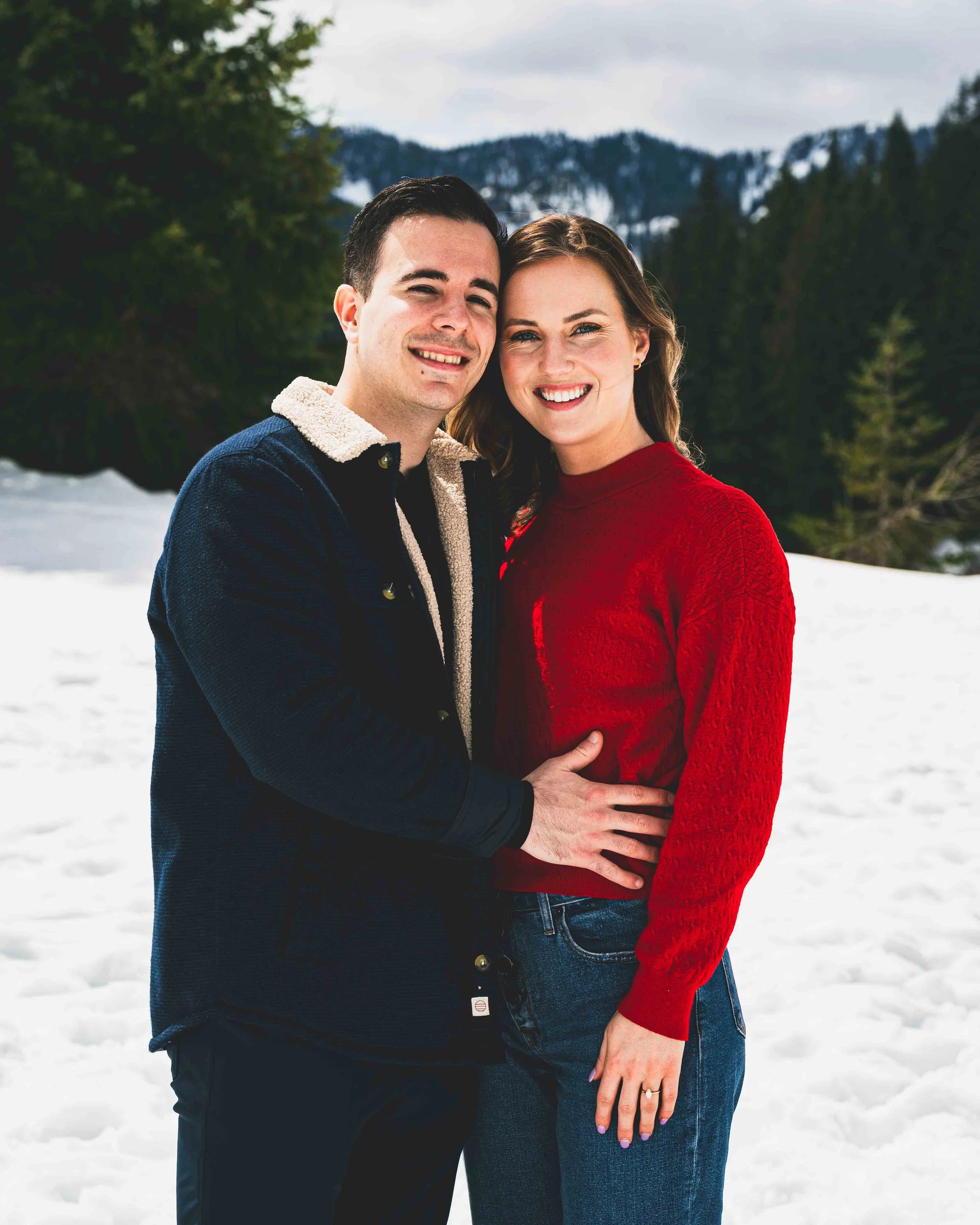 A couple smiling and standing close together outdoors in a snowy mountainous landscape, with forested mountains in the background. The man is wearing a navy jacket with a shearling collar, and the woman is wearing a red sweater and blue jeans.