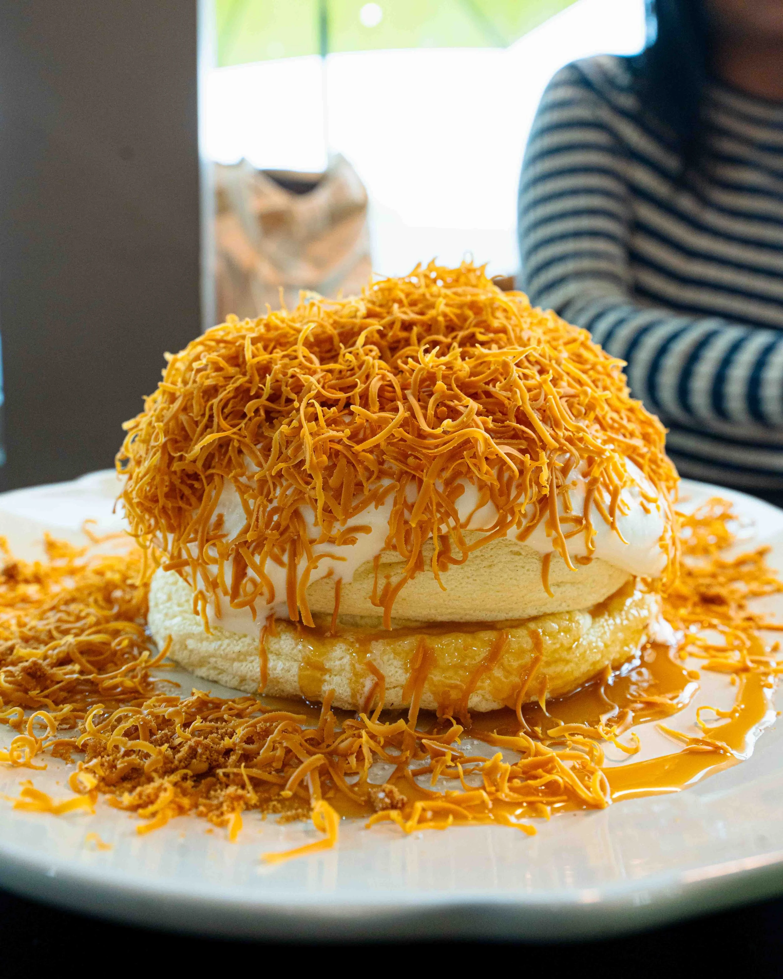 Close-up of a dessert with shredded cheese on top, with a table seen in the background and a person wearing a striped shirt partially visible.
