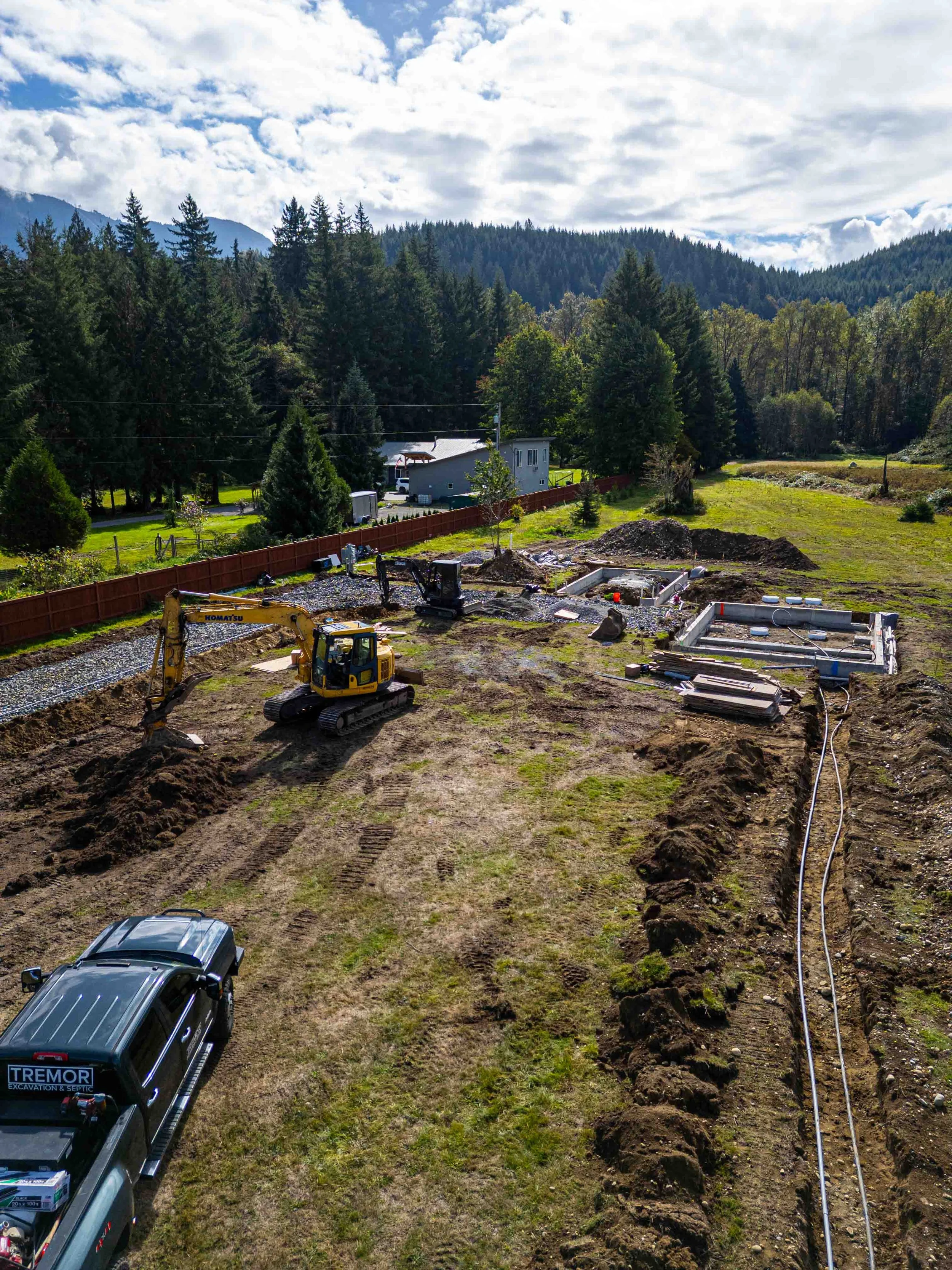 Construction site with excavator, utility trenches, and vehicles in a grassy field surrounded by trees and mountains under a partly cloudy sky.