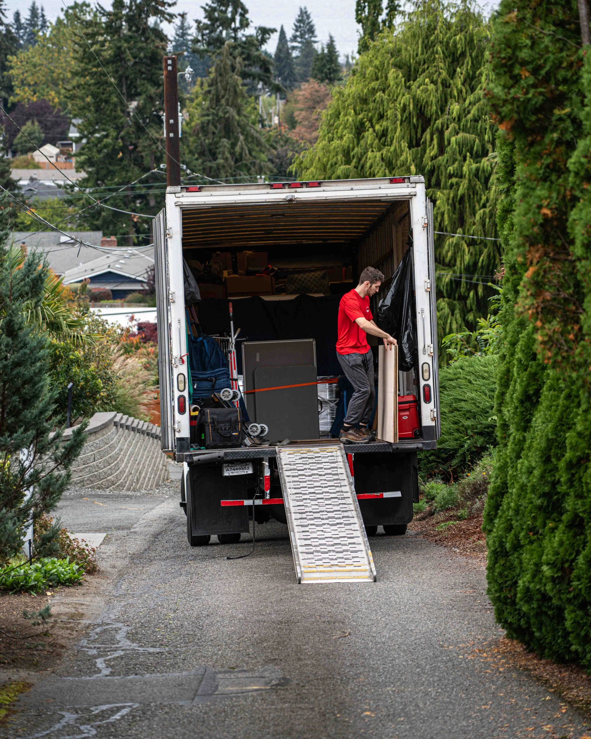 A man in a red shirt and gray pants loading objects into a moving truck in a suburban neighborhood with greenery and trees.