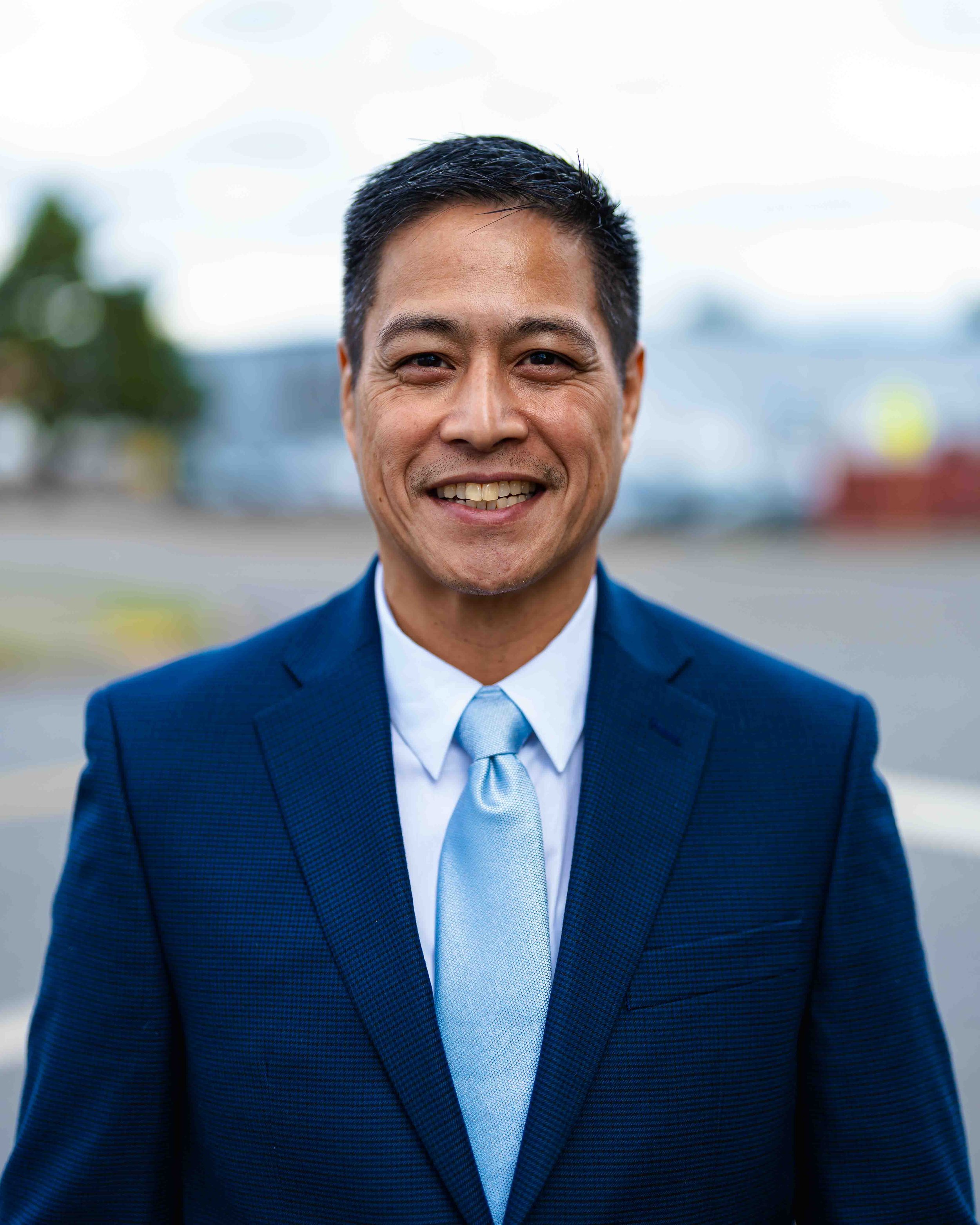 A smiling man in a blue suit, white shirt, and light blue tie, standing outdoors with blurred trees and sky in the background.
