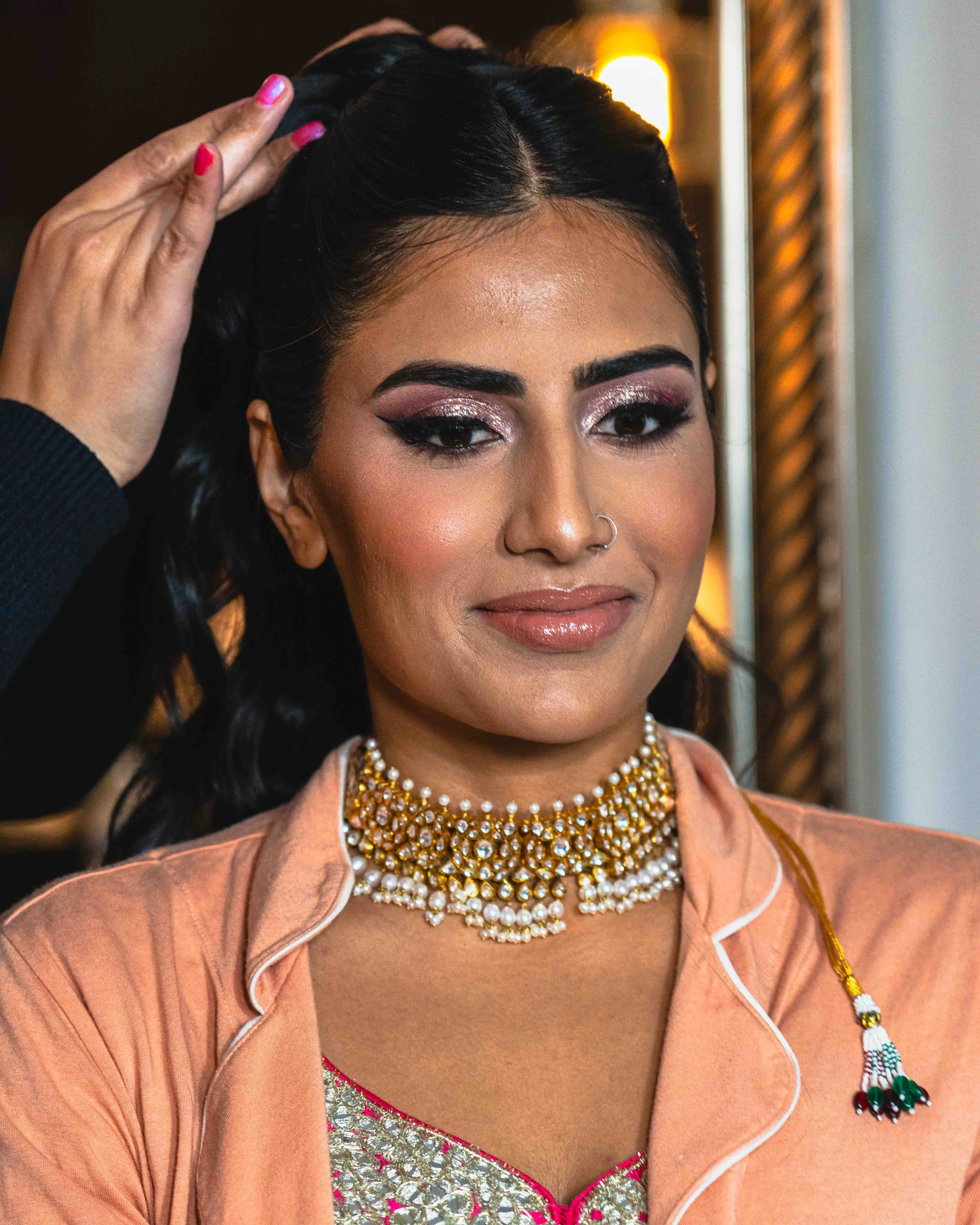 Close-up of a woman with makeup, wearing a gold and pearl necklace, a peach-colored traditional dress with pink embroidery, and a peach dupatta. Someone adjusts her hair in a dimly lit setting.