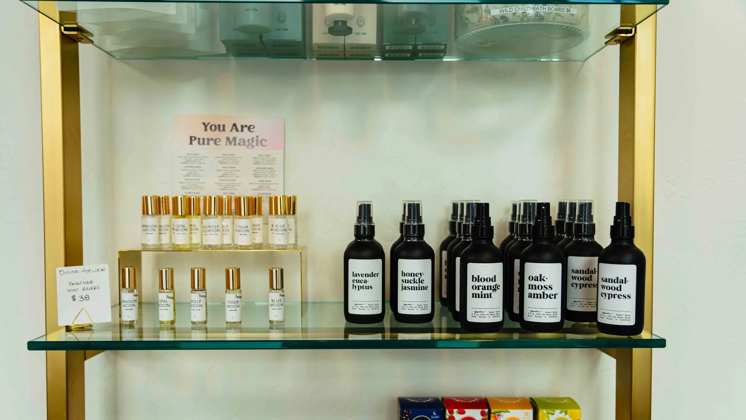 A display shelf with small perfume bottles labeled with scent names like lavender eucalyptus, honey-suckle jasmine, blood orange mint, oak-moss, sandalwood, and cypress. A sign reads "You Are Pure Magic." There is a price tag for mini perfume rollers