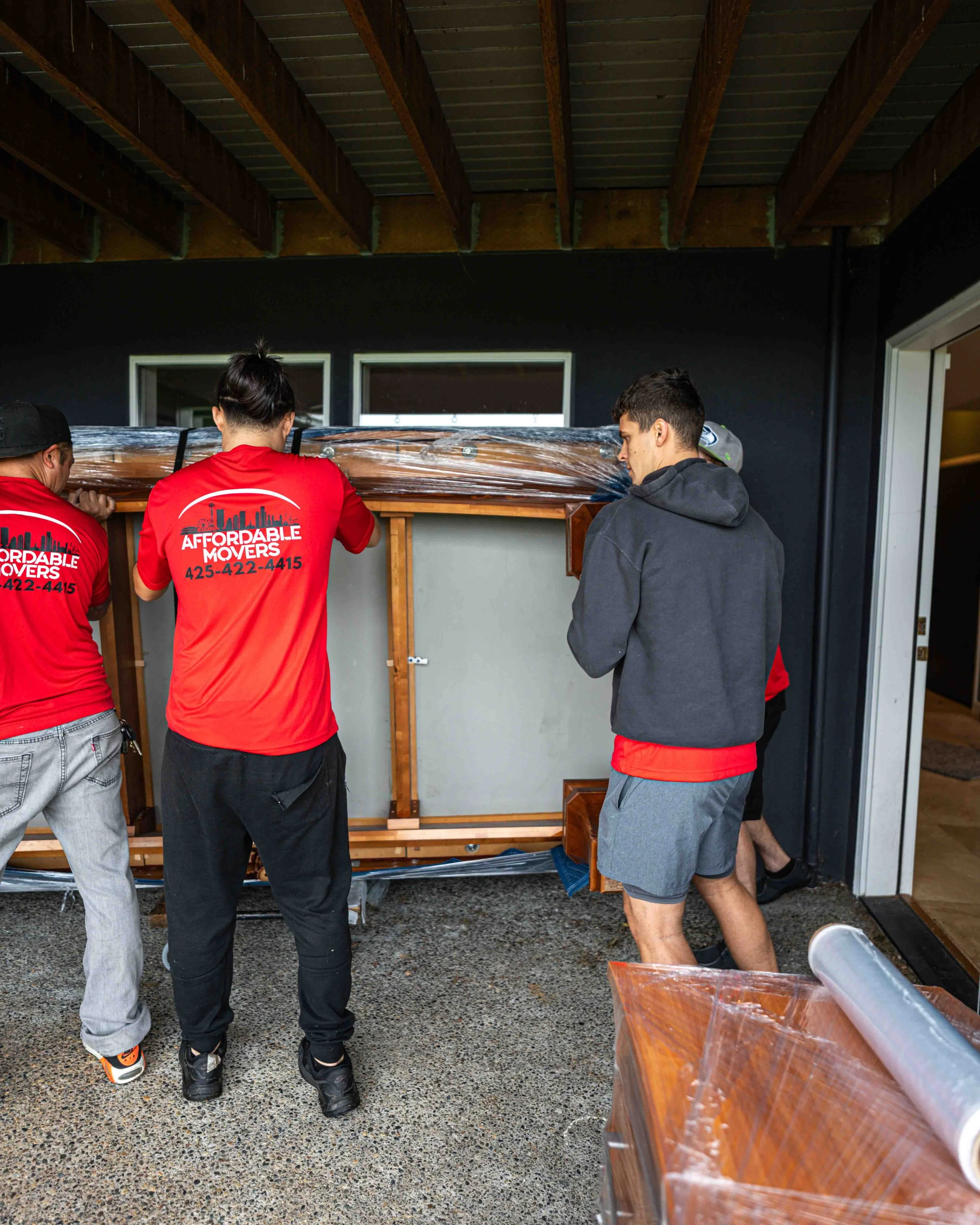 Threemen lifting a wooden piece wrapped in plastic, using a black strap at a construction site.