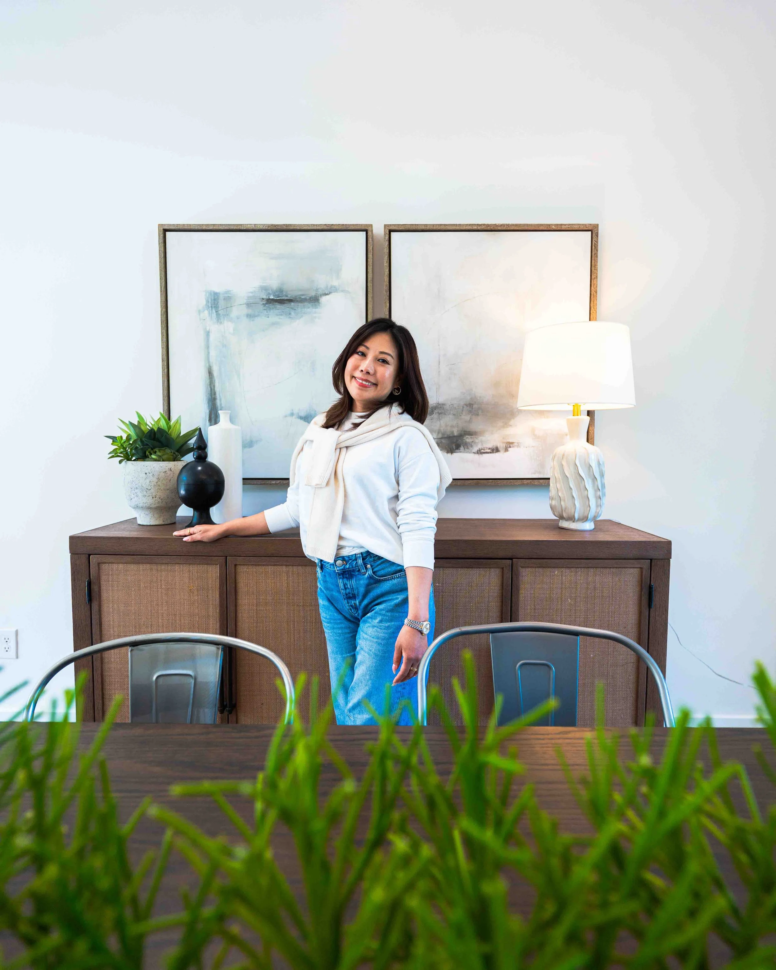 A woman standing in front of a wooden dining table, with a green plant and a black vase, behind her wall decorations, a table lamp, and abstract artwork.