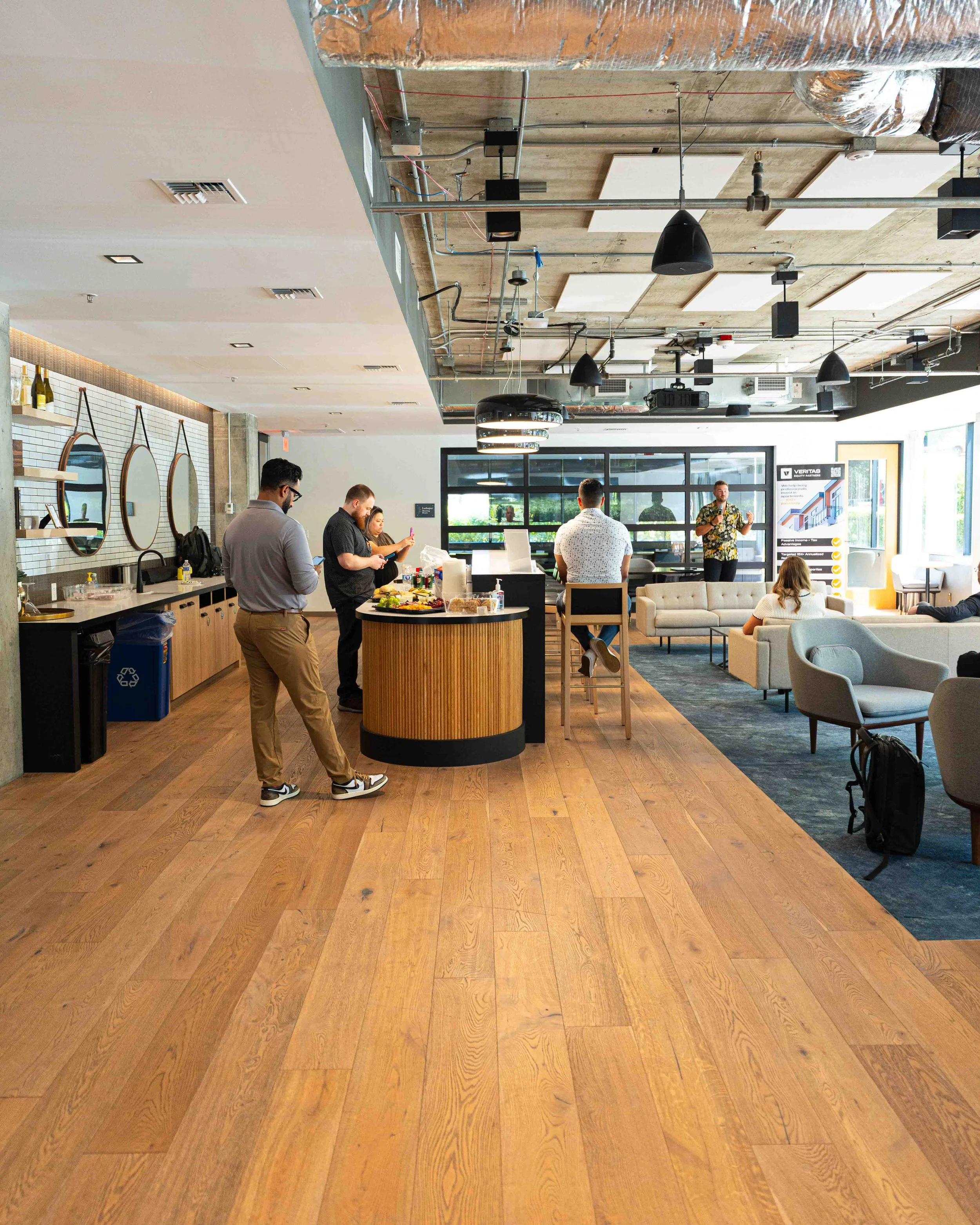 People standing and sitting in a modern, open-concept office space with a wooden floor, a decorated wall with round mirrors, and an exposed ceiling with various light fixtures and ductwork. One person appears to be giving a presentation.