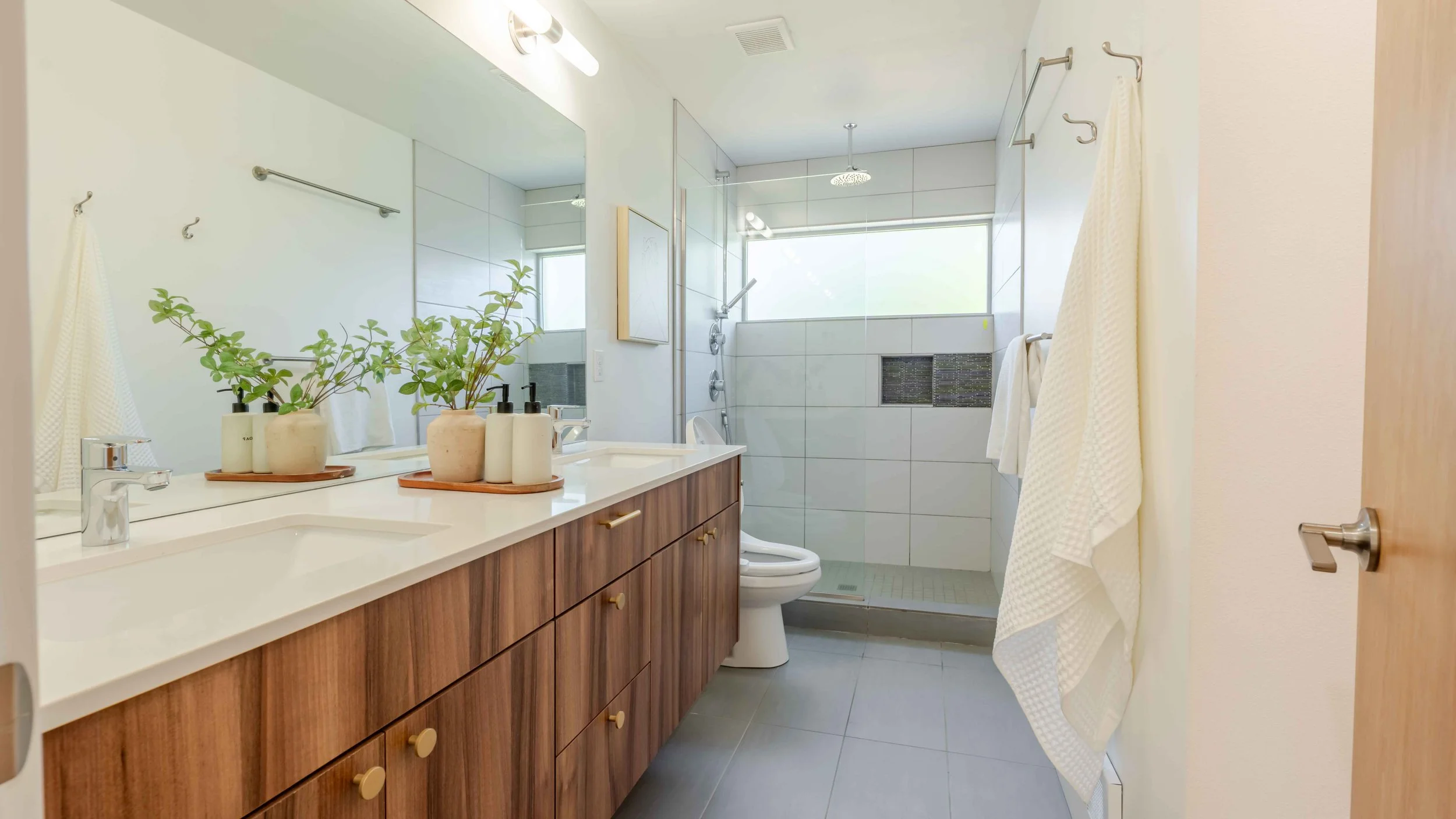 Bathroom with a wooden vanity, plants, bottles, a white countertop, a mirror, a shower area with a rain showerhead, a window, and a toilet.