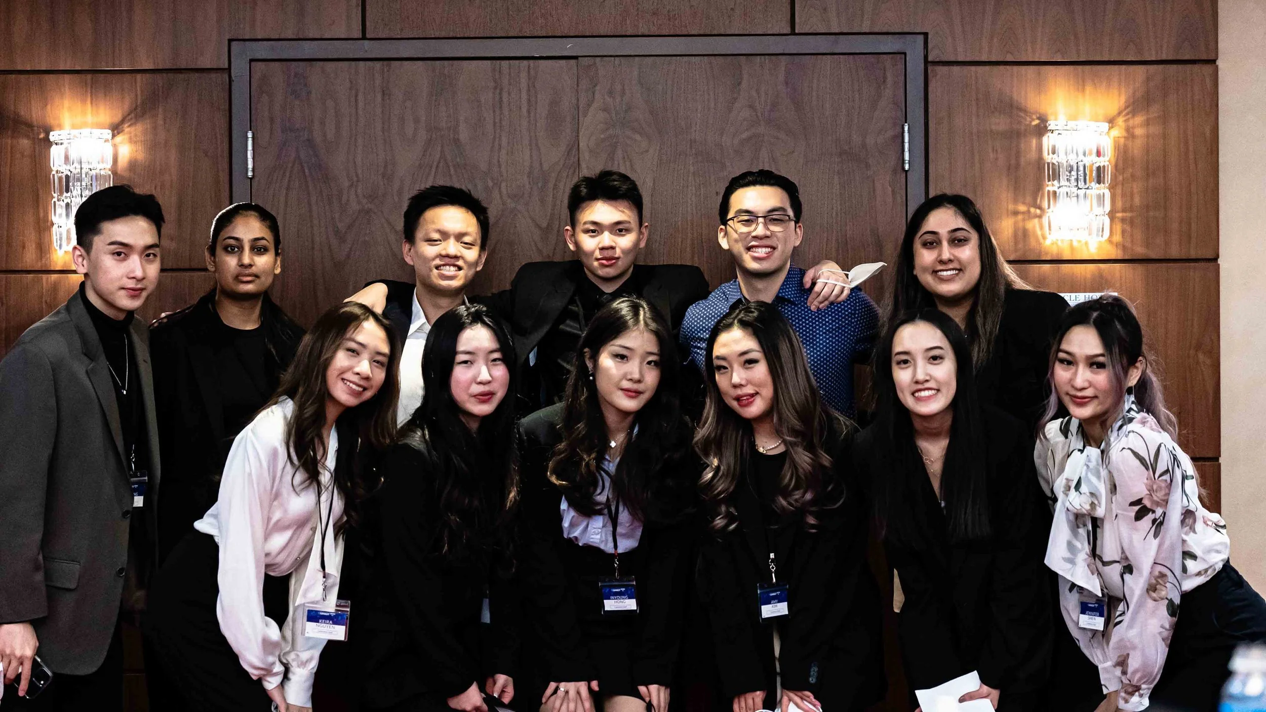 Group of young adults posing for a photo at an indoor event, with a wooden wall and wall-mounted lights in the background.