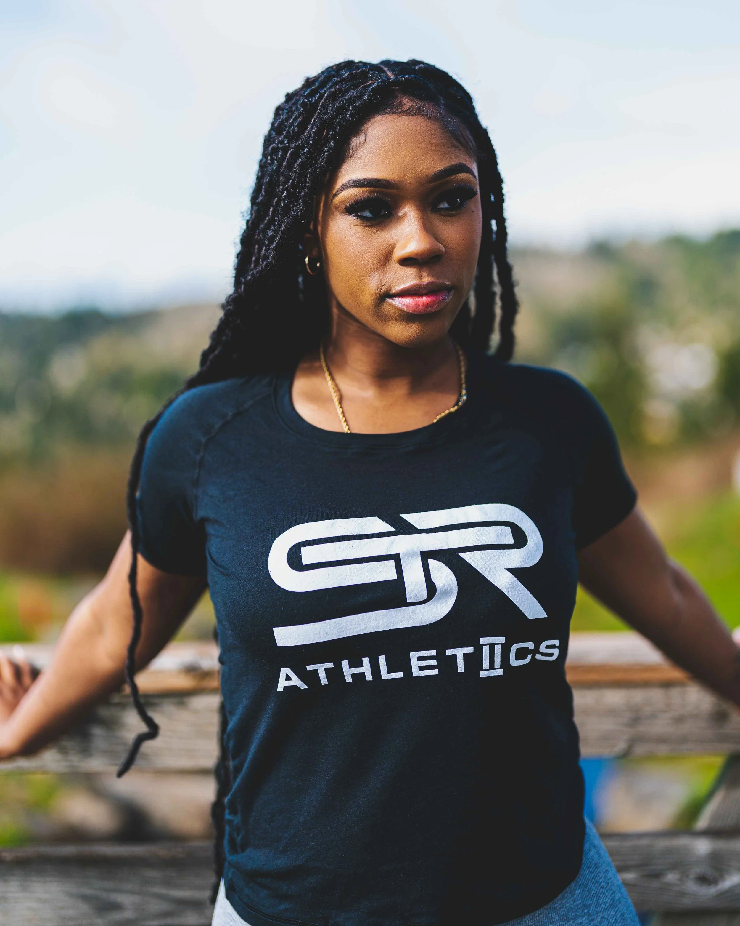 Close-up of a woman with long dreadlocks wearing a black athletic T-shirt with white text, outdoors with a blurred background of trees and sky.