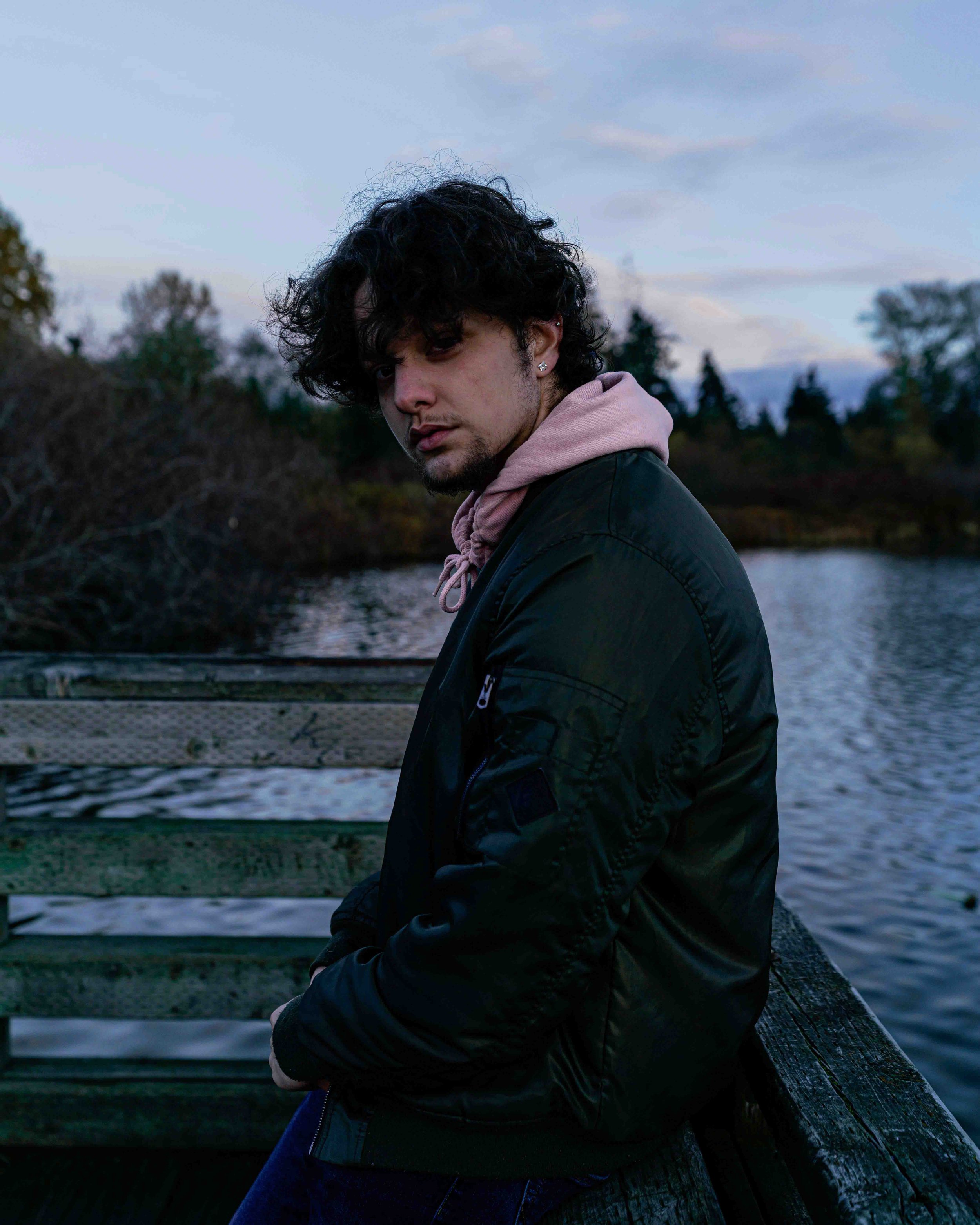 A young man with curly dark hair and earrings, wearing a pink hoodie under a black jacket, sitting on a wooden dock by a river or lake at dusk, with trees and clouds in the background.