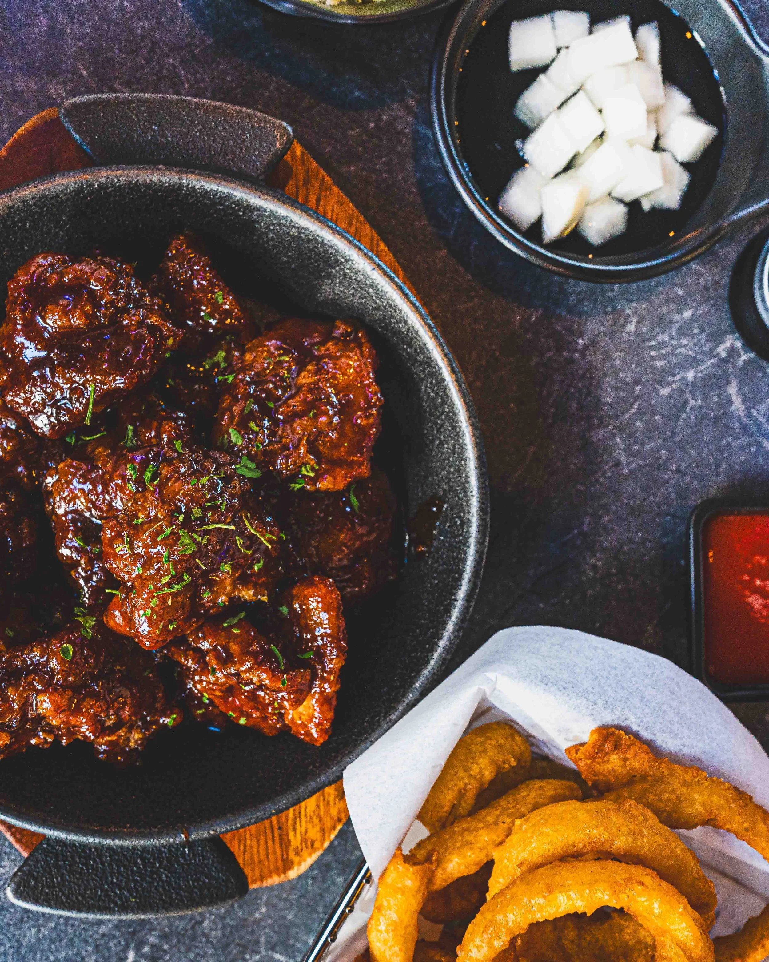 Bowl of Korean fried chicken wings covered in sauce with chopped green herbs, nearby a black bowl of diced white radish, and a basket of onion rings and fried chicken strips.