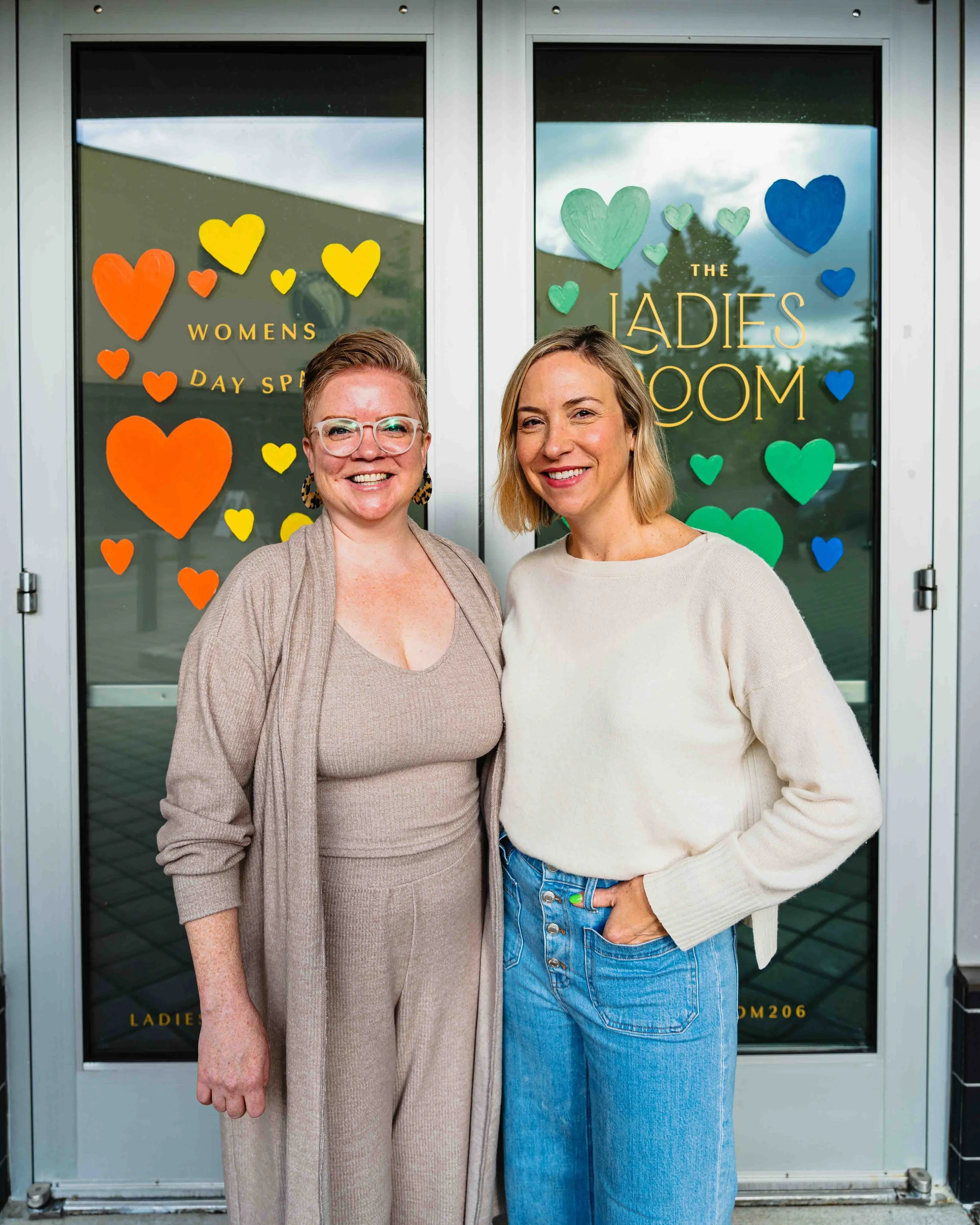 Two women standing in front of a glass door with colorful heart decorations and gold lettering, smiling at the camera.