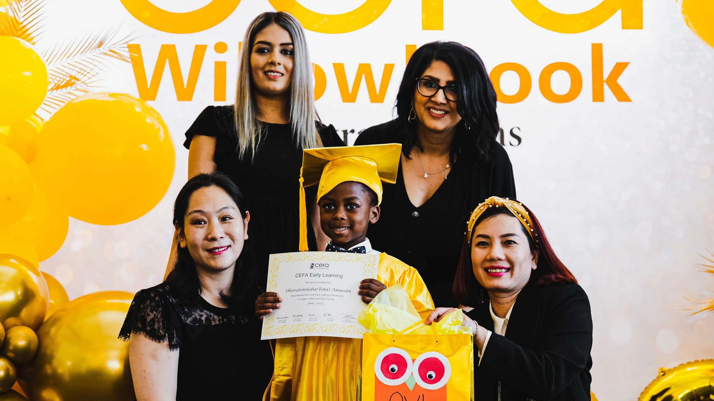 Group of women and a young graduate in a yellow cap and gown holding a diploma, celebrating graduation at a cheerful event with yellow balloons and decorations.