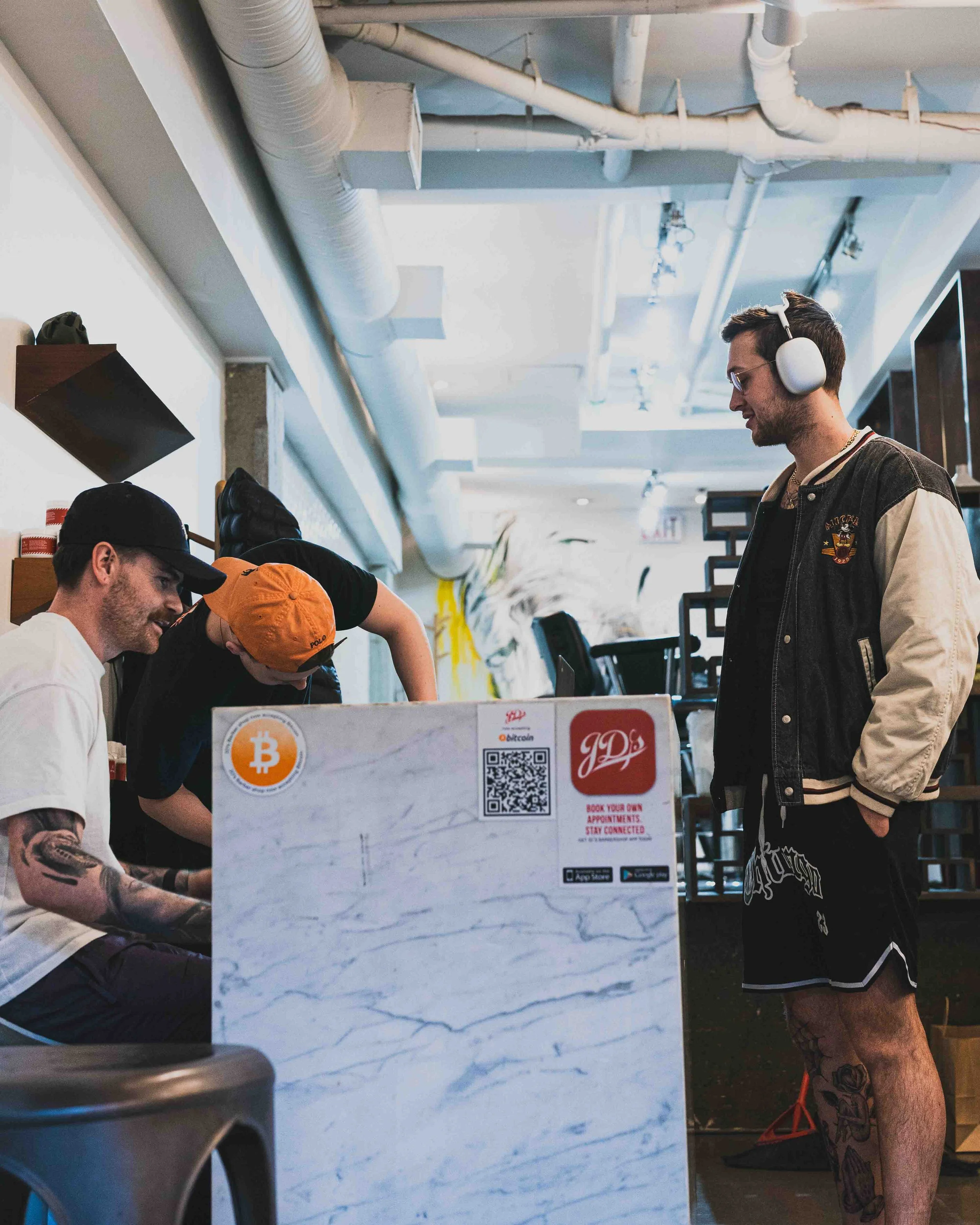 A young man with glasses, a black varsity jacket with patches, and headphones, standing at a counter, smiling and talking to a person wearing an orange cap, inside a modern café or restaurant with an industrial ceiling. There is a marble-like counter