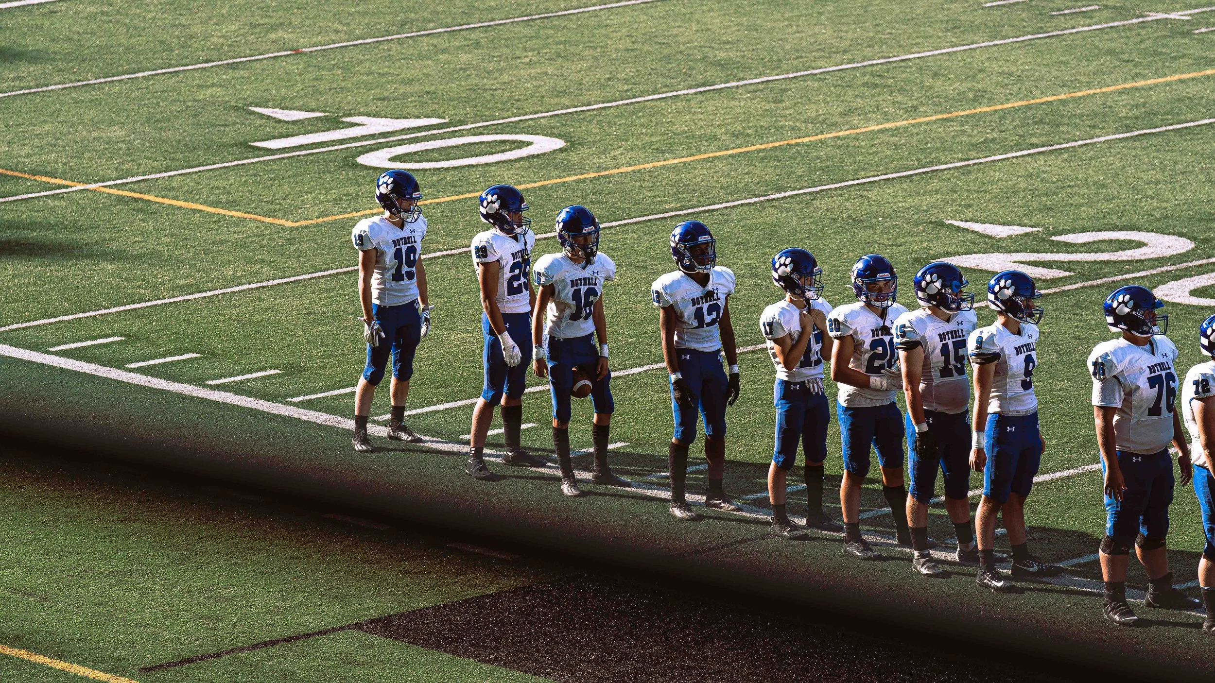 A line of football players in uniform standing on a field before a game, with the number 20 visible on some of their jerseys.