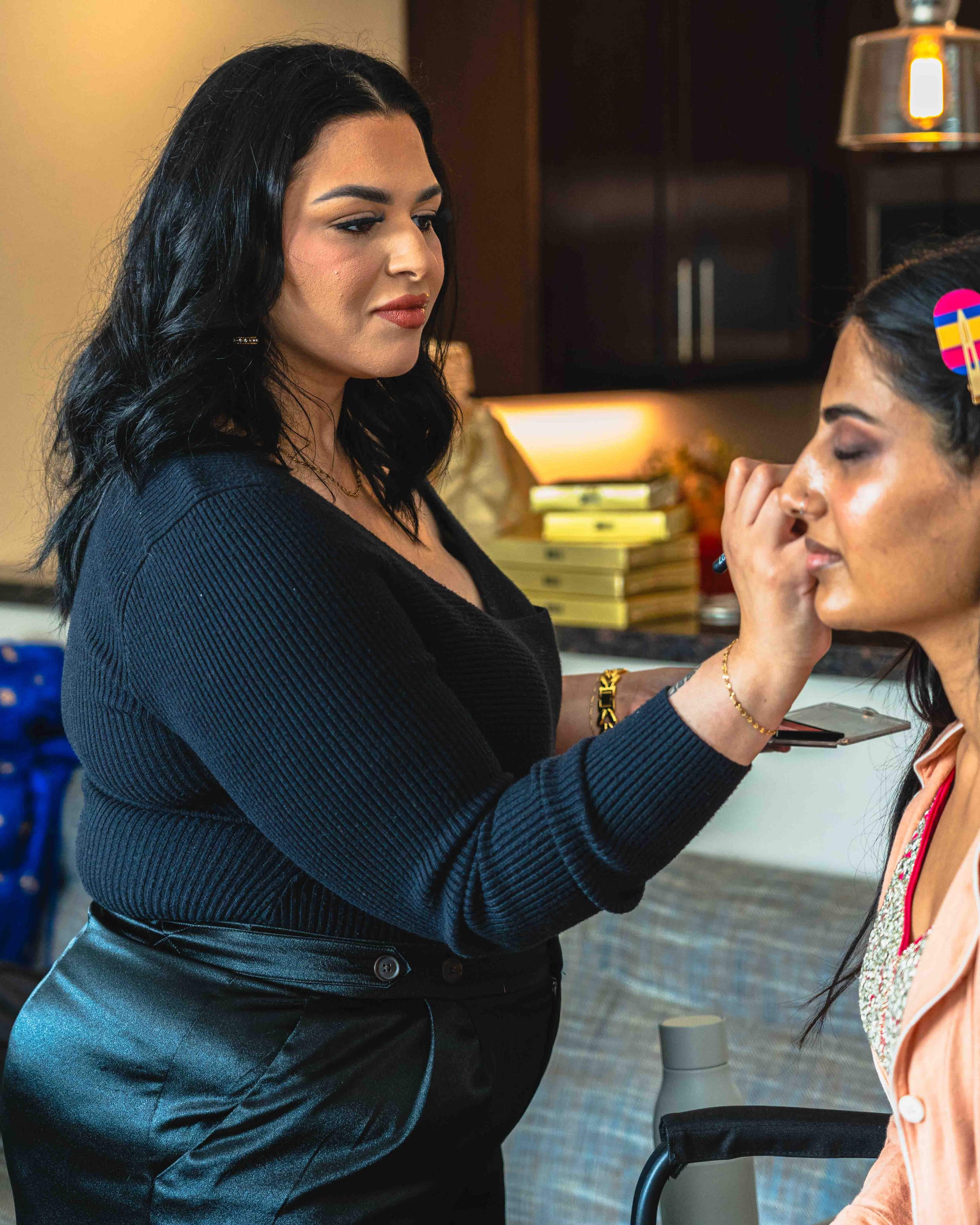 A woman with black hair and light skin applying makeup to another woman with darker skin and black hair, who is seated with her eyes closed and hair clipped back, in a room with warm lighting, a stack of books, and a gray speaker in the background.