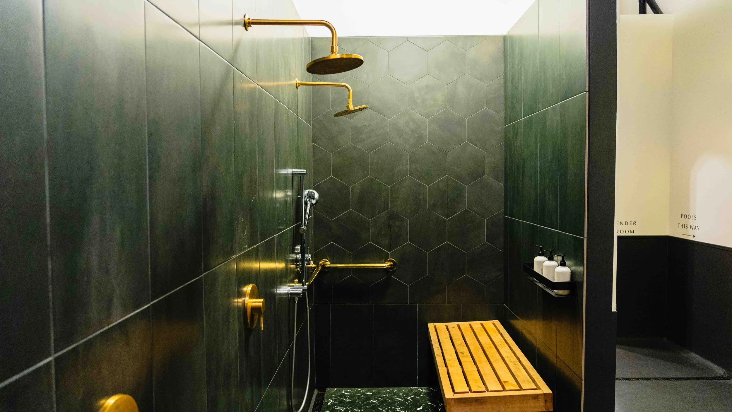 A modern shower with green hexagonal tiles on the wall, featuring gold shower fixtures, a handheld shower head, a wooden bench, and a black shelf with toiletries, and a sign indicating the direction to the pool room.