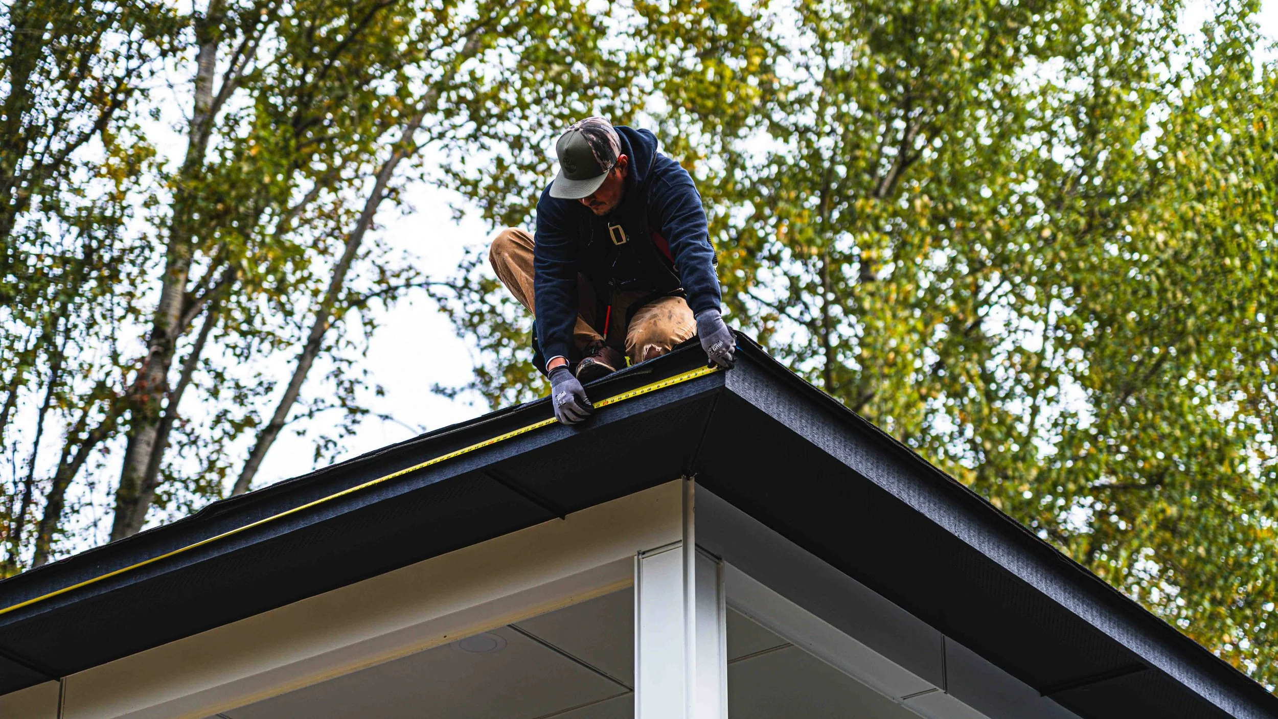 A person installing or repairing a roof on a house with a tape measure, outdoors among trees.