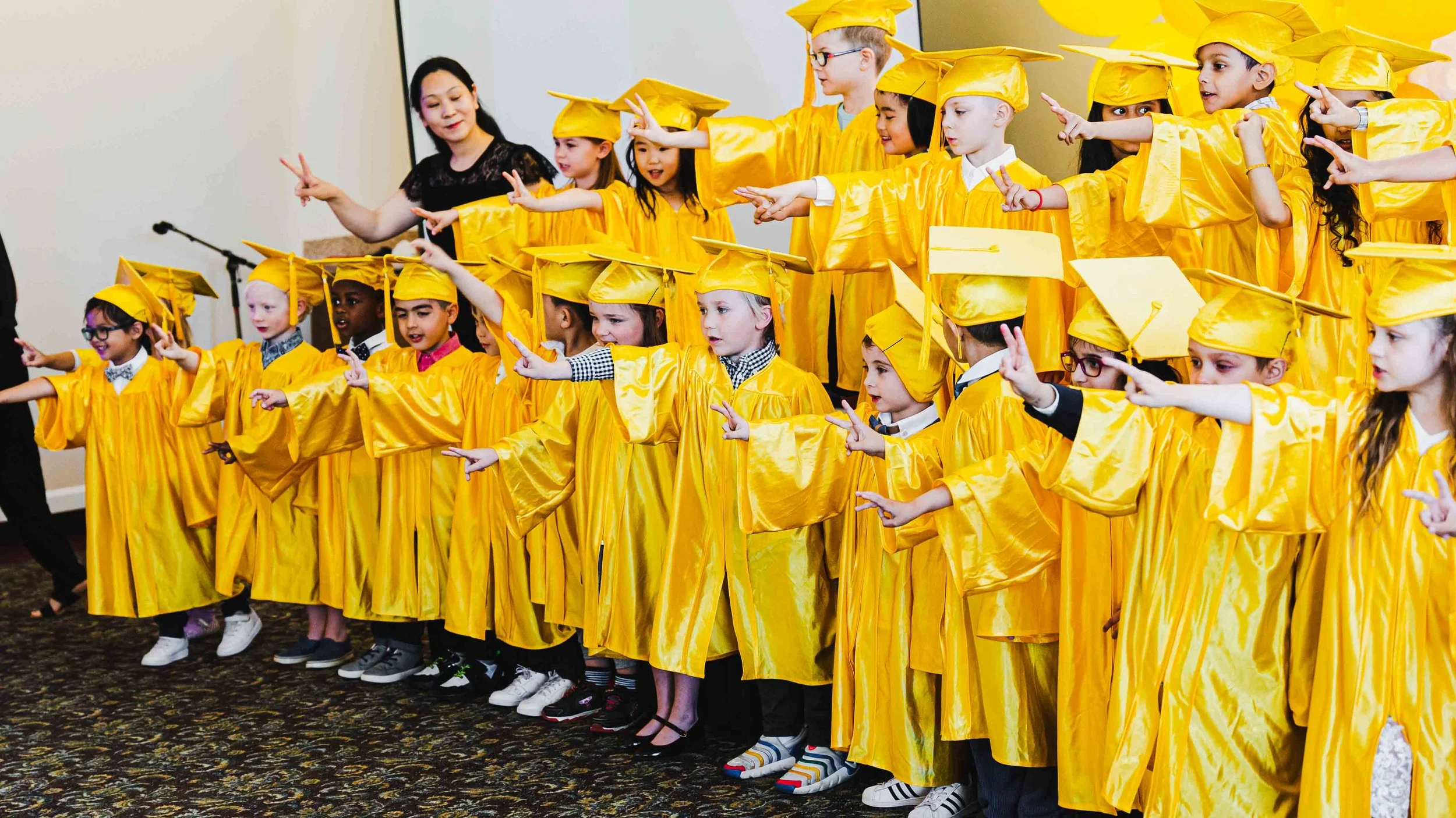 Children wearing yellow graduation gowns and caps stand in a line, raising their arms and pointing in a direction, during a graduation ceremony.