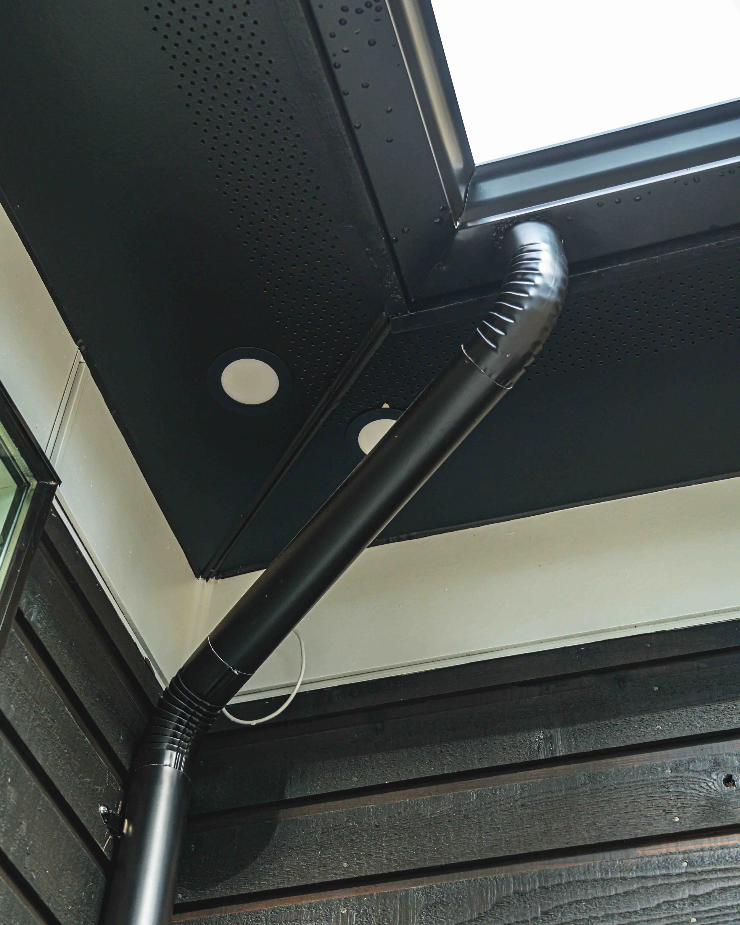 Close-up of a black ceiling with a black duct pipe, circular lights, and perforated panels.