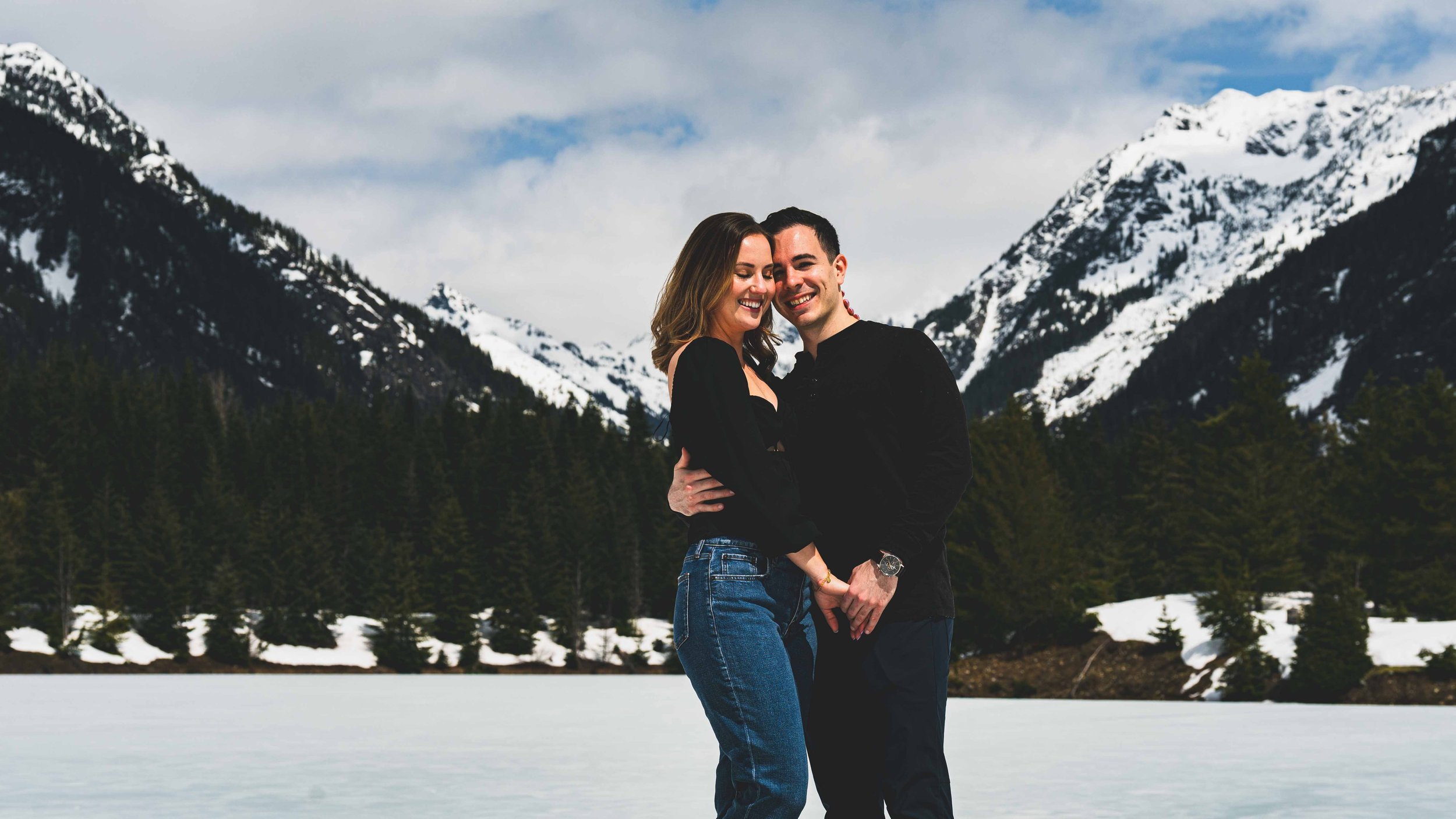 A couple stands near a frozen lake with snow-capped mountains and evergreen trees in the background, hugging and smiling.