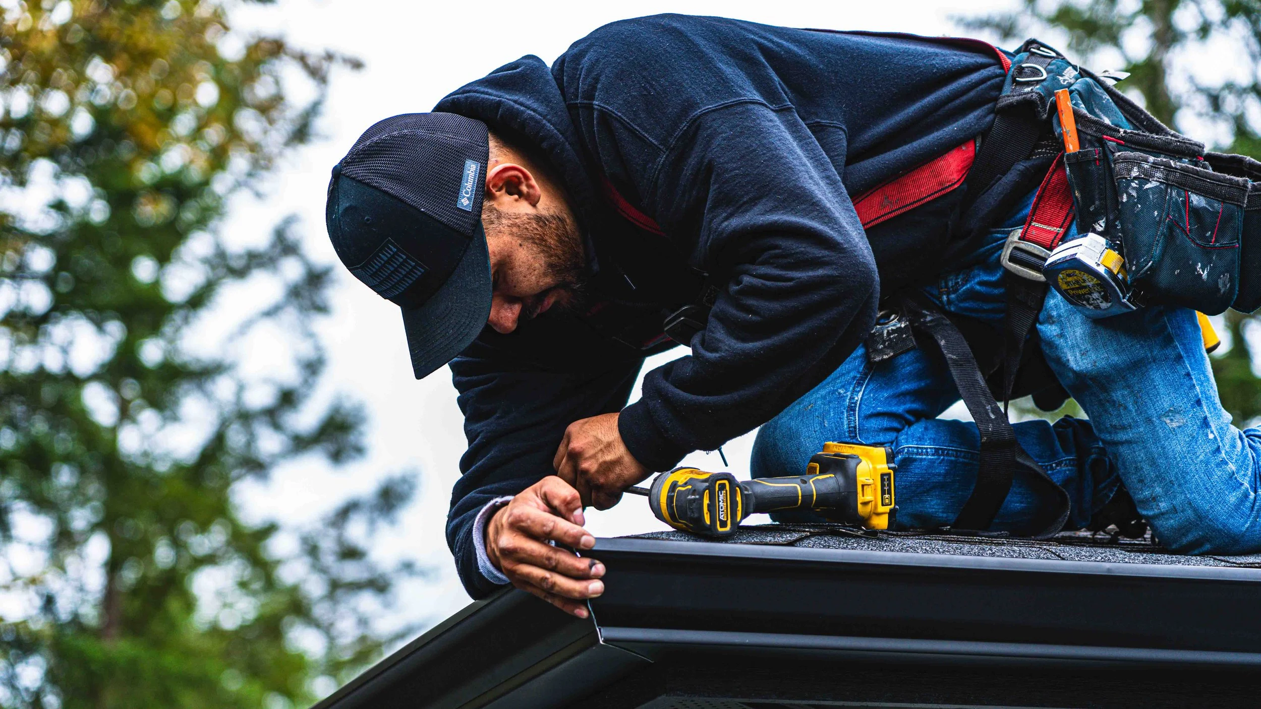 Man working on a roof using a yellow power drill, wearing a black hat, black hoodie, blue jeans, and a safety harness, with trees in the background.