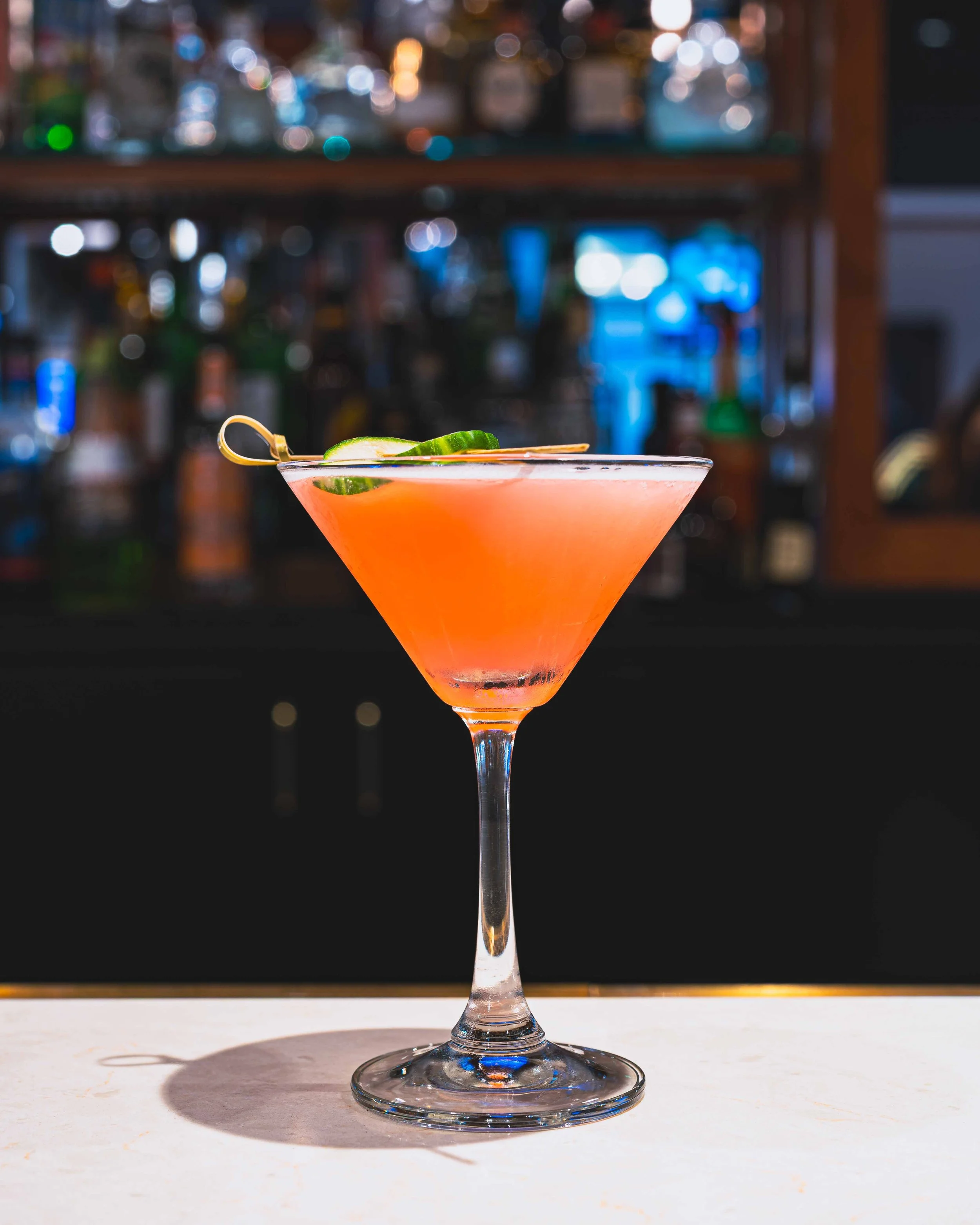 A pink cocktail in a martini glass garnished with cucumber slices, placed on a white surface with a blurred bar background.