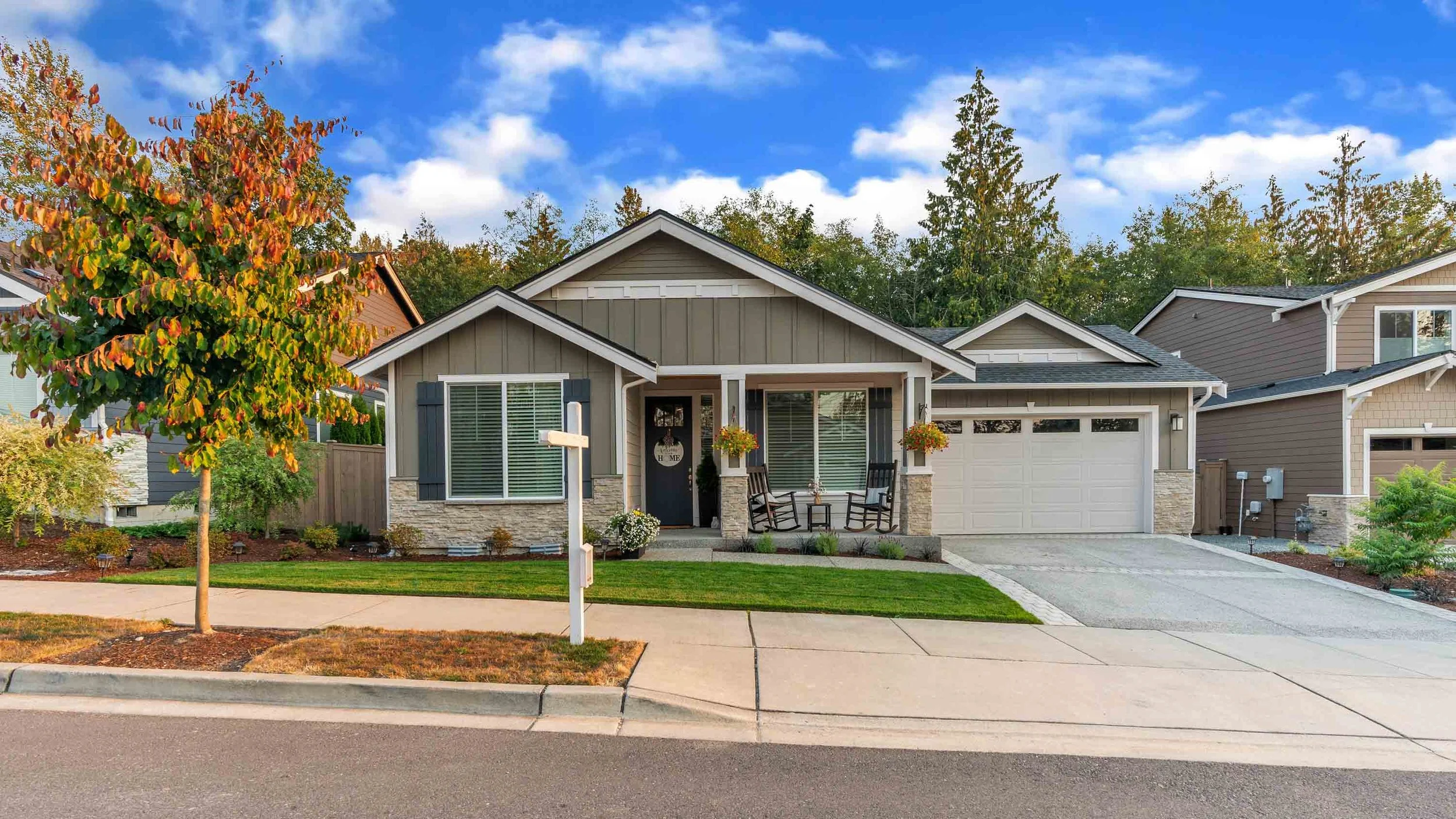 A modern suburban house with a front porch, rocking chairs, and hanging flower baskets, surrounded by a well-maintained lawn and trees with autumn foliage. The sky is partly cloudy.