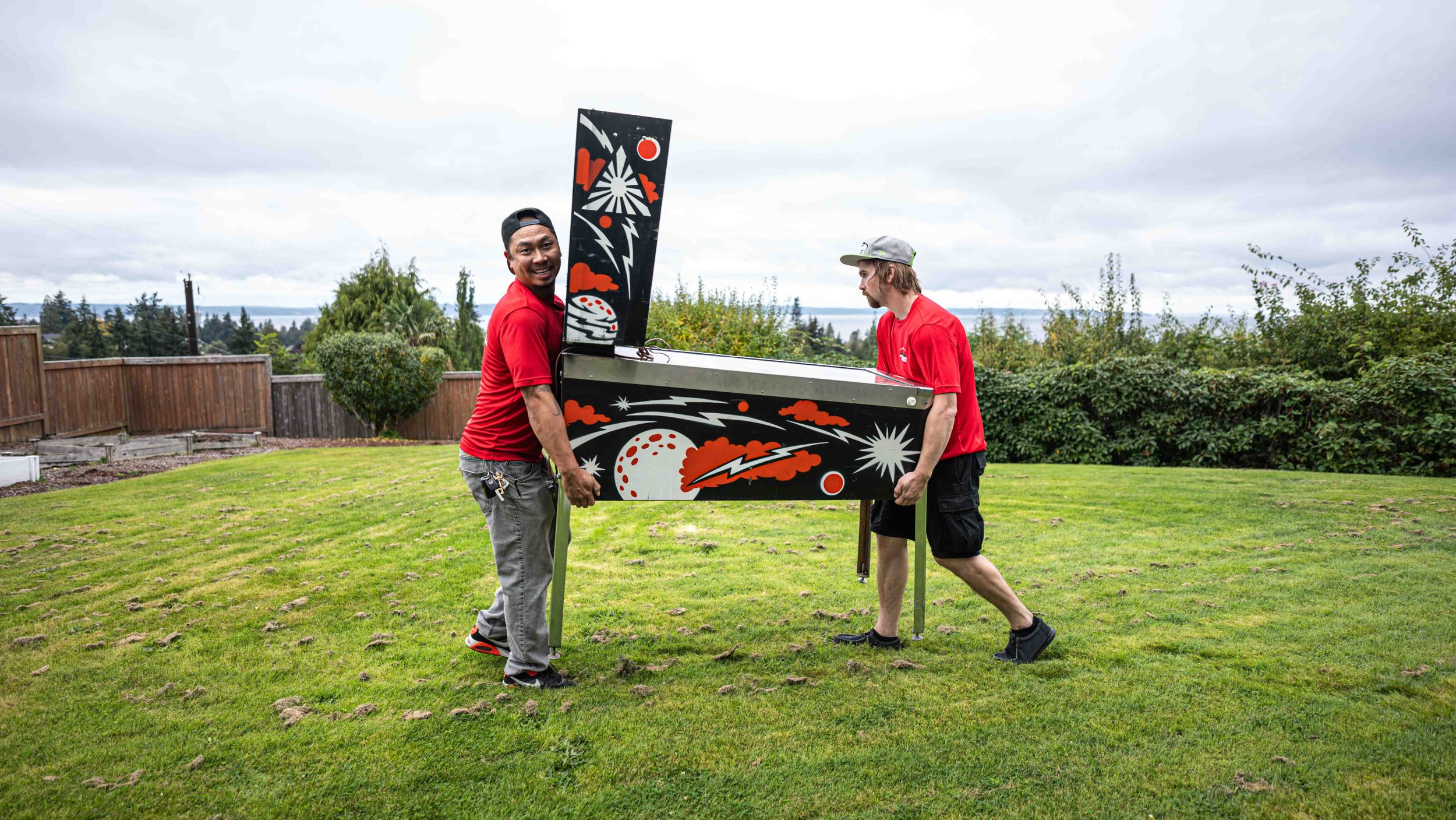 Two men in red shirts assembling a pinball machine outdoors. The pinball machine has a space-themed design with planets, clouds, lightning bolts, and stars. One man is smiling at the camera while holding the machine, and the other is focused on the a