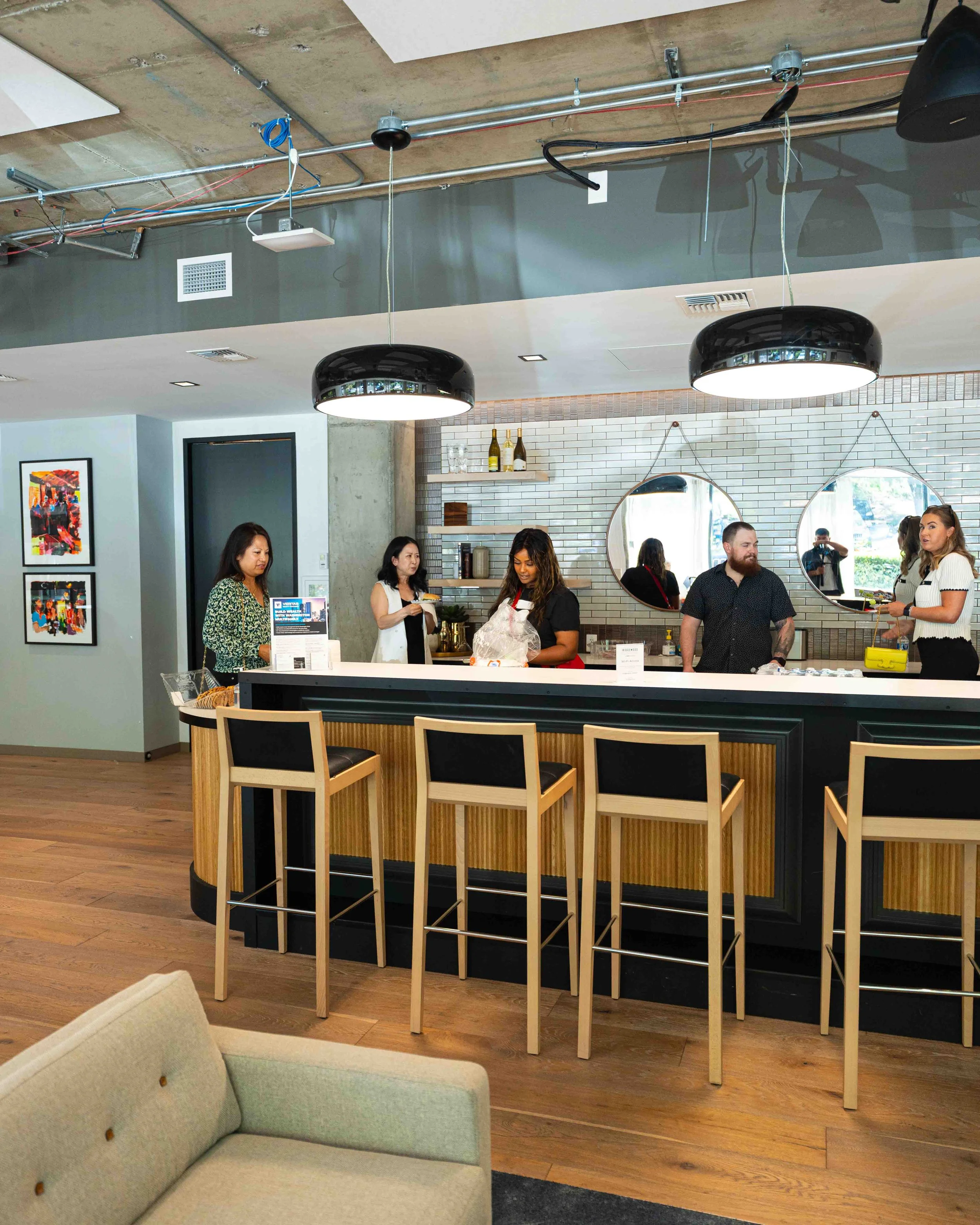 Interior of a restaurant or cafe with a modern design. The ceiling has exposed pipes and lighting fixtures. There are several people standing and talking behind a counter. The wall behind has two round mirrors and a shelf with bottles. In the foregro