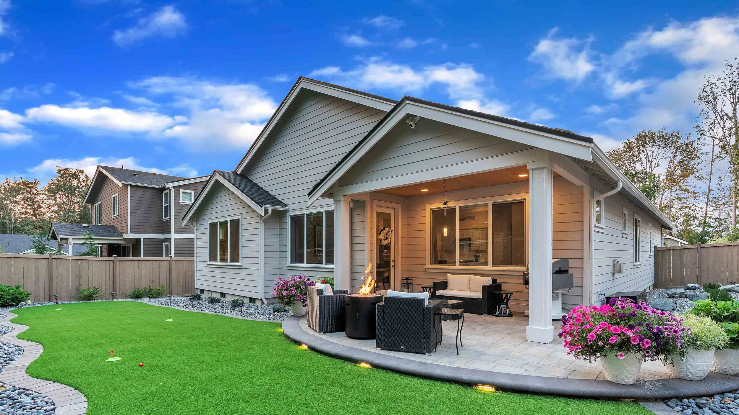 Backyard patio with outdoor furniture, a fire pit, potted flowers, and a well-maintained lawn, with a house and a fence in the background.