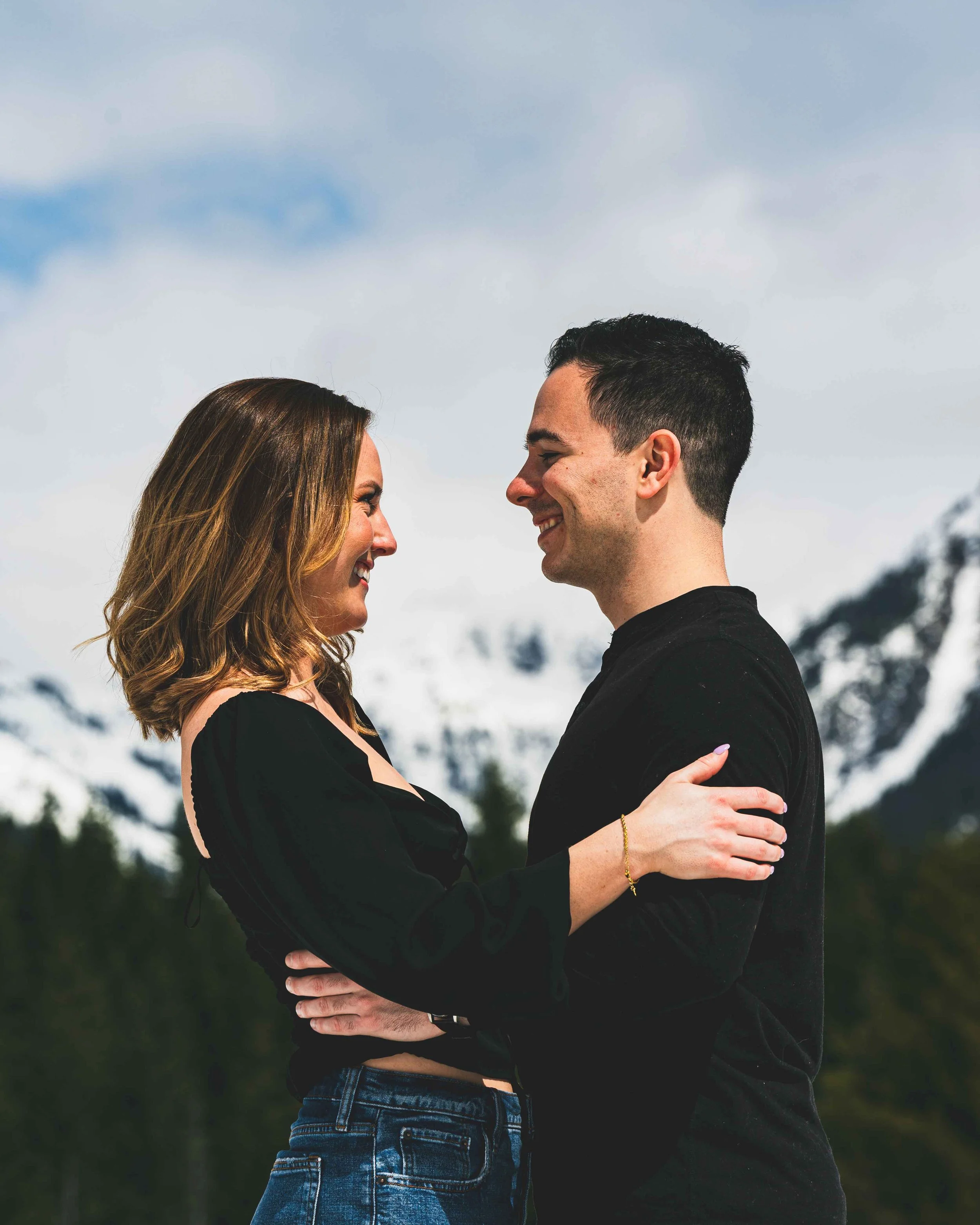 A couple is standing close together outdoors with snowy mountains in the background, smiling and looking into each other's eyes. They are holding each other affectionately.