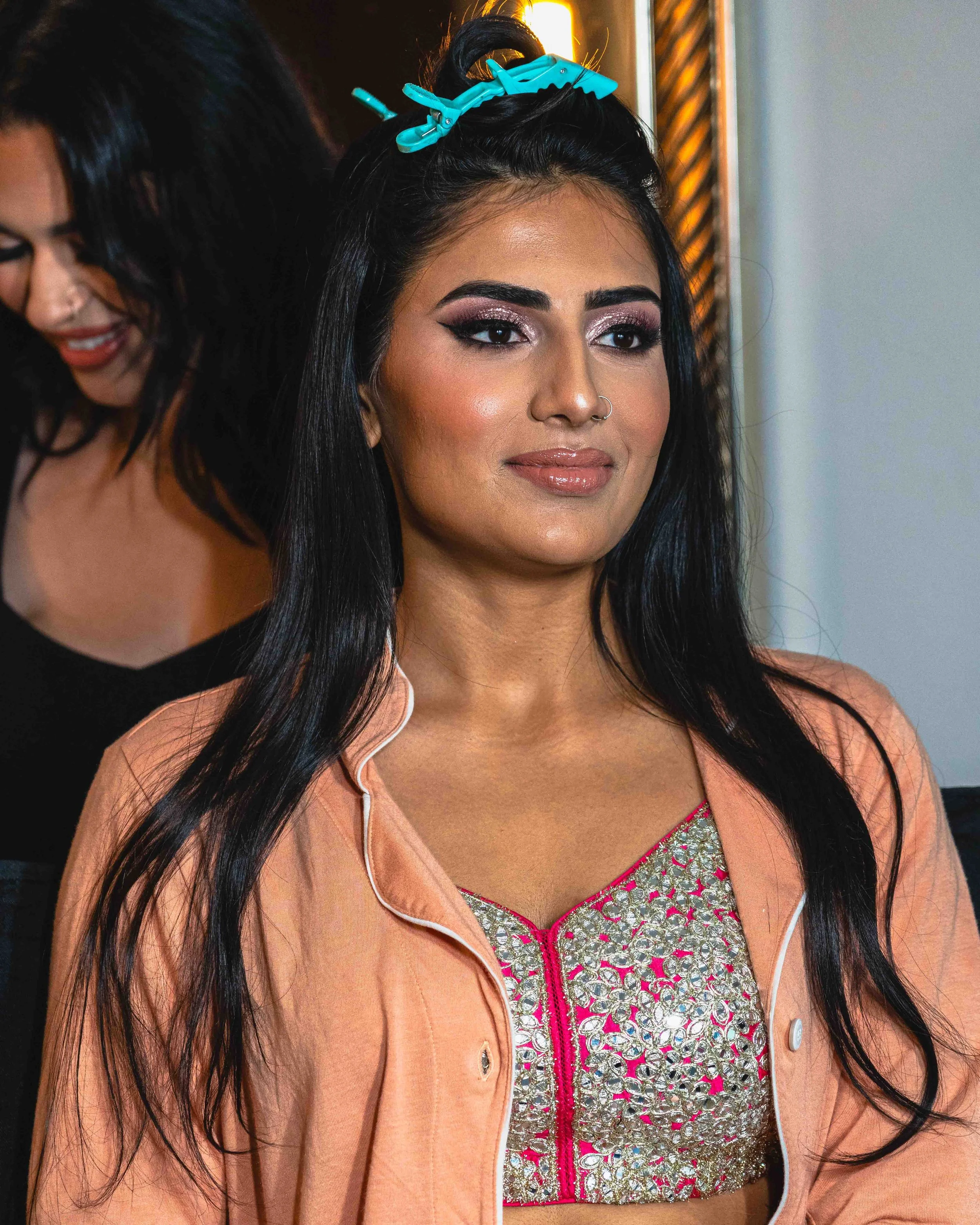A woman with long black hair and makeup, wearing a peach-colored jacket with a ruffled collar, over a pink and silver embroidered dress, at an indoor event.