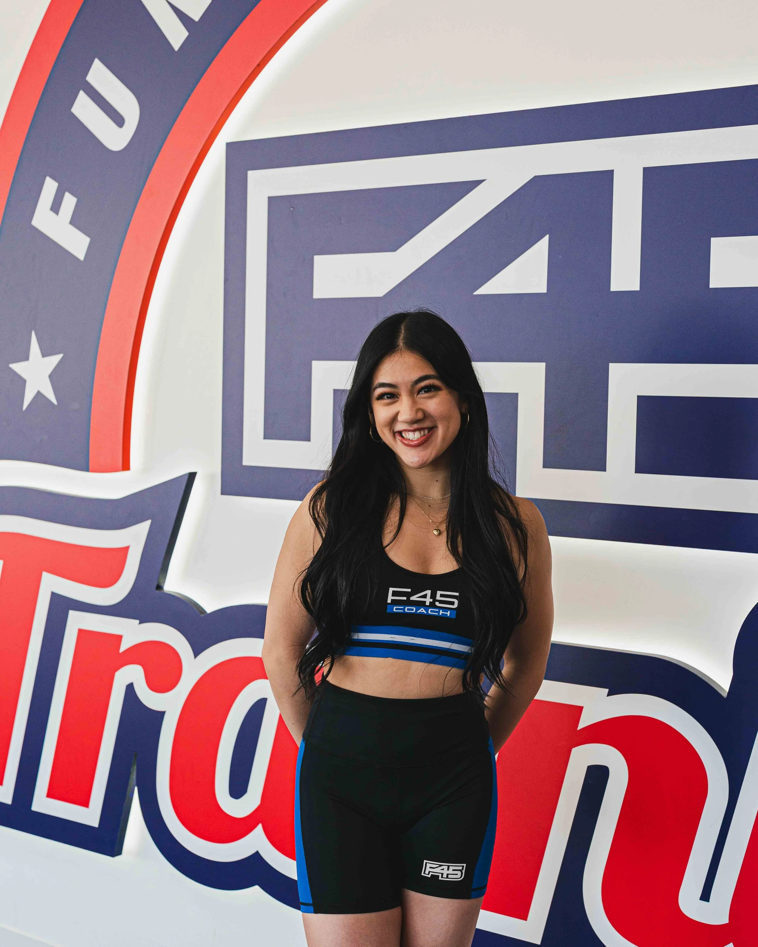 A young woman smiling, wearing a black crop top with blue and white accents and black shorts with a logo, stands in front of a large patriotic sign with red, white, and blue colors.