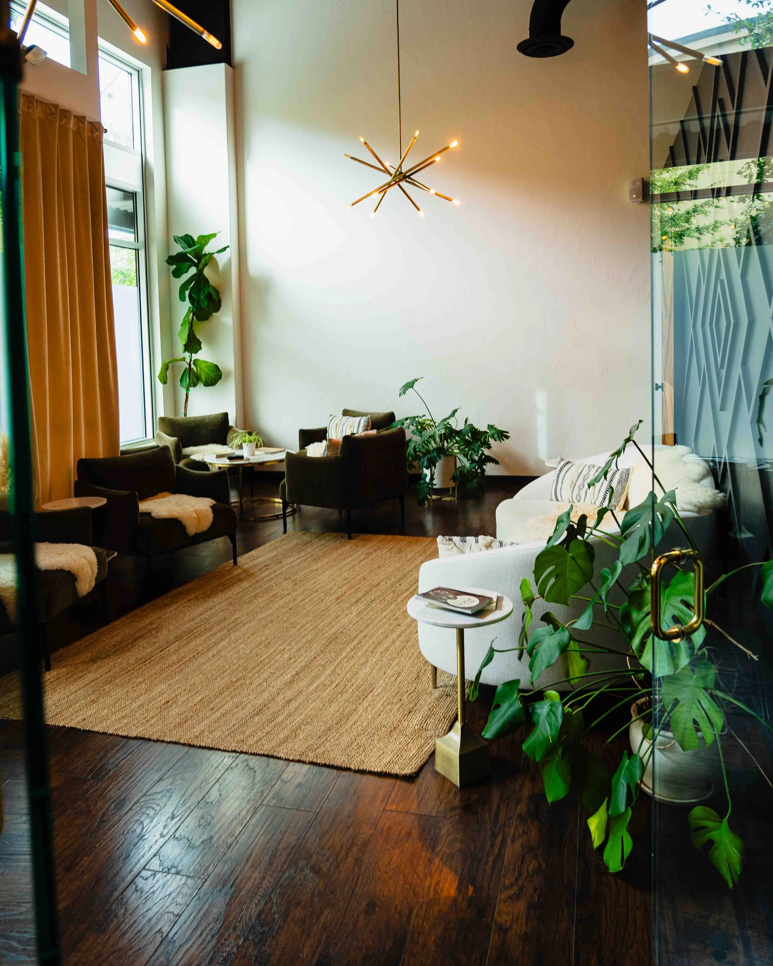 Interior of a modern living room with a high ceiling, large windows, wooden floors, a beige rug, green plants, a white sofa with patterned pillows, dark chairs arranged around a table, and a contemporary chandelier hanging from the ceiling.